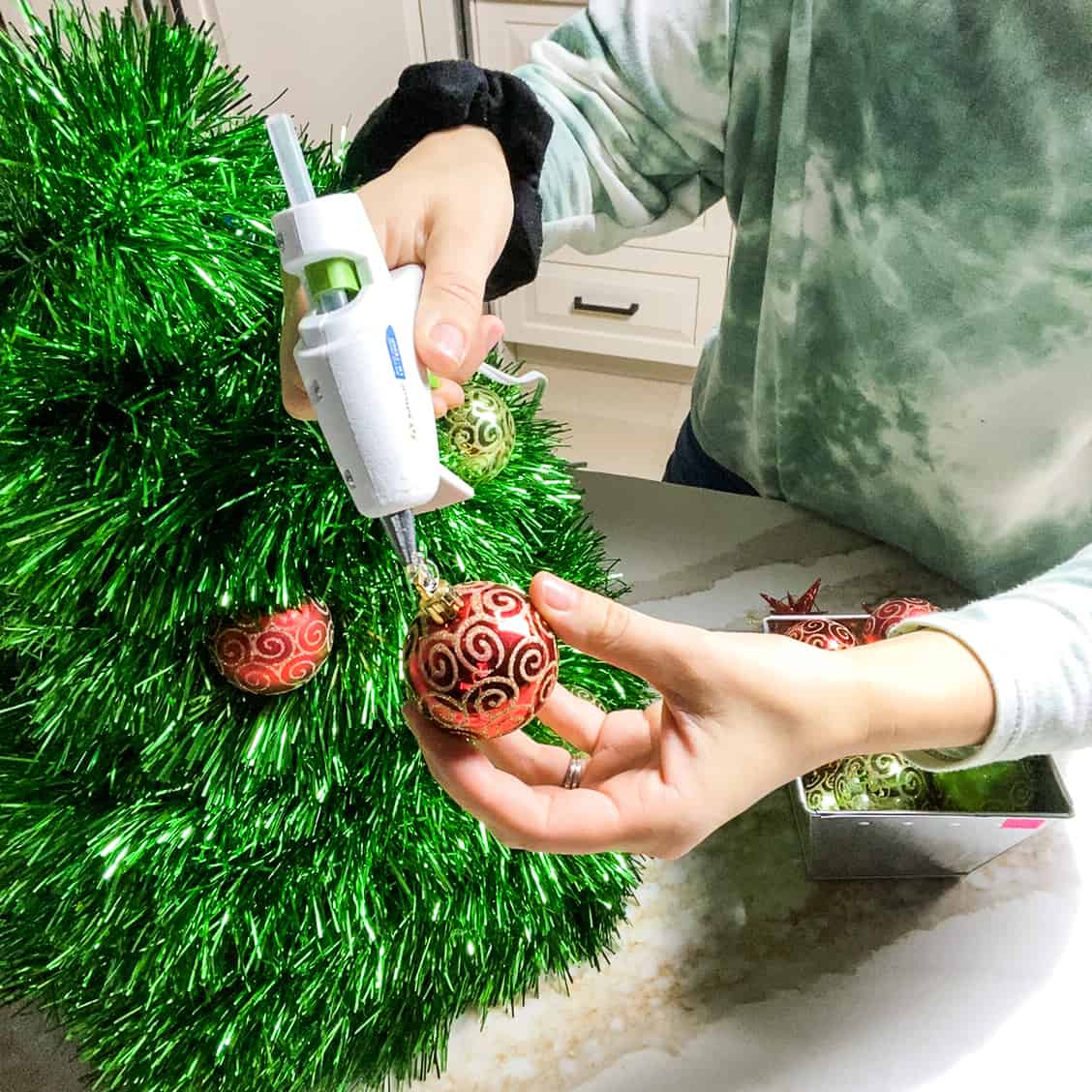 Squeezing hot glue onto the end of a Christmas ball decoration so that it can be permanently attached to the wire hanger Christmas tree.