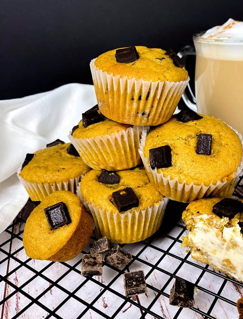 A pyramid of pumpkin muffins with large chocolate chunks on the top and a frothy latte lurking in the foreground. Black background with a black cooling rack and white linen napkin underneath.