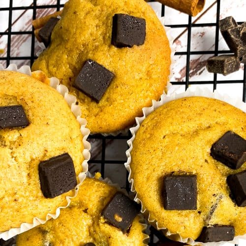 Pumpkin chocolate muffins on a black wire rack with chocolate chunks and cinnamon sticks.