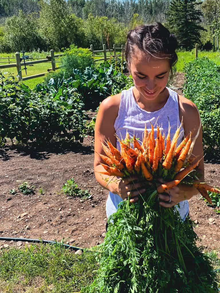 Woman holding a bouquet of carrots in front of her green garden.