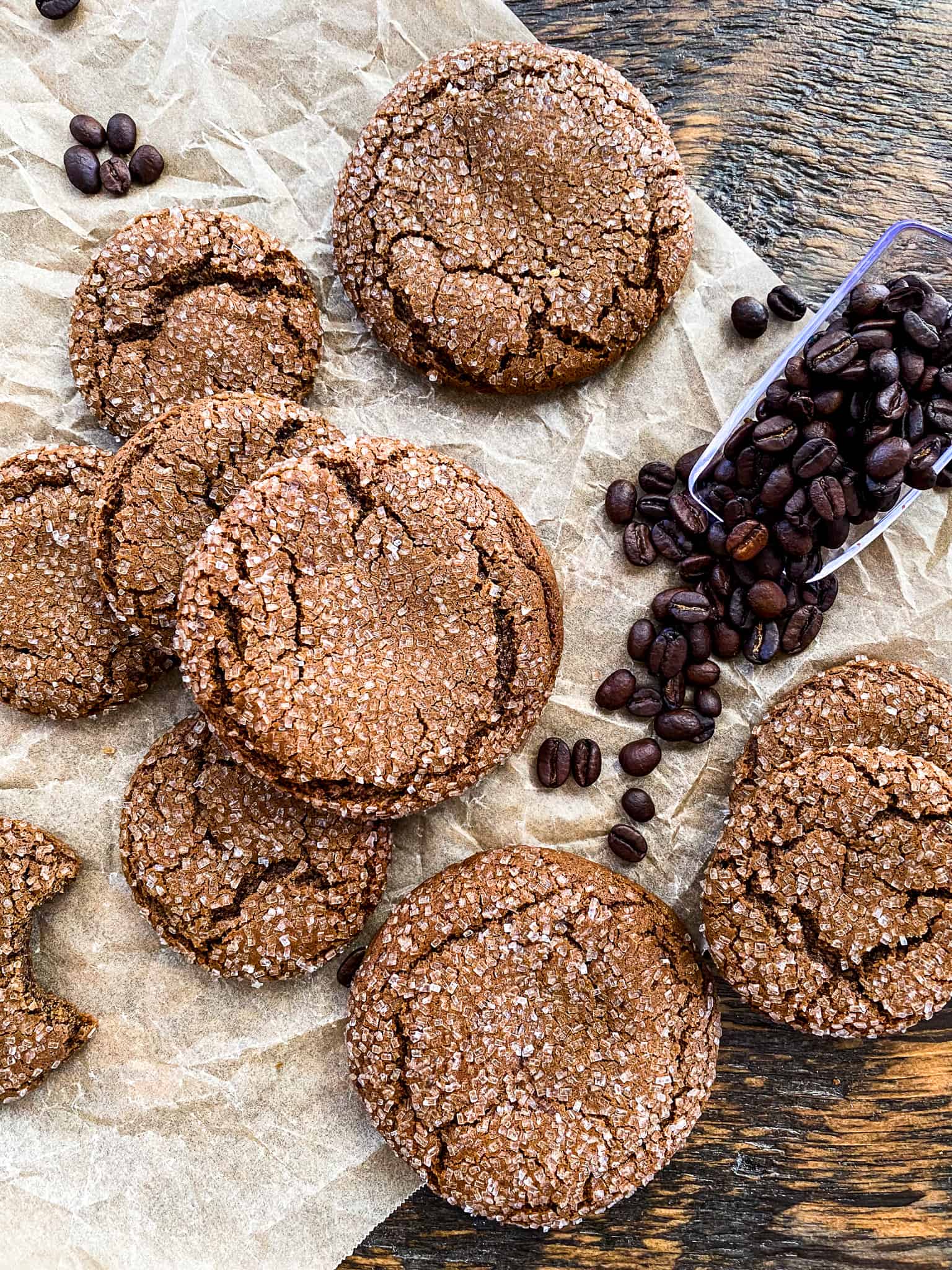 Overhead view of espresso cookies with coffee beans spilling out onto parchment paper. Lots of big cookies coated in fancy sugar, boasting large cracked tops and moist insides.