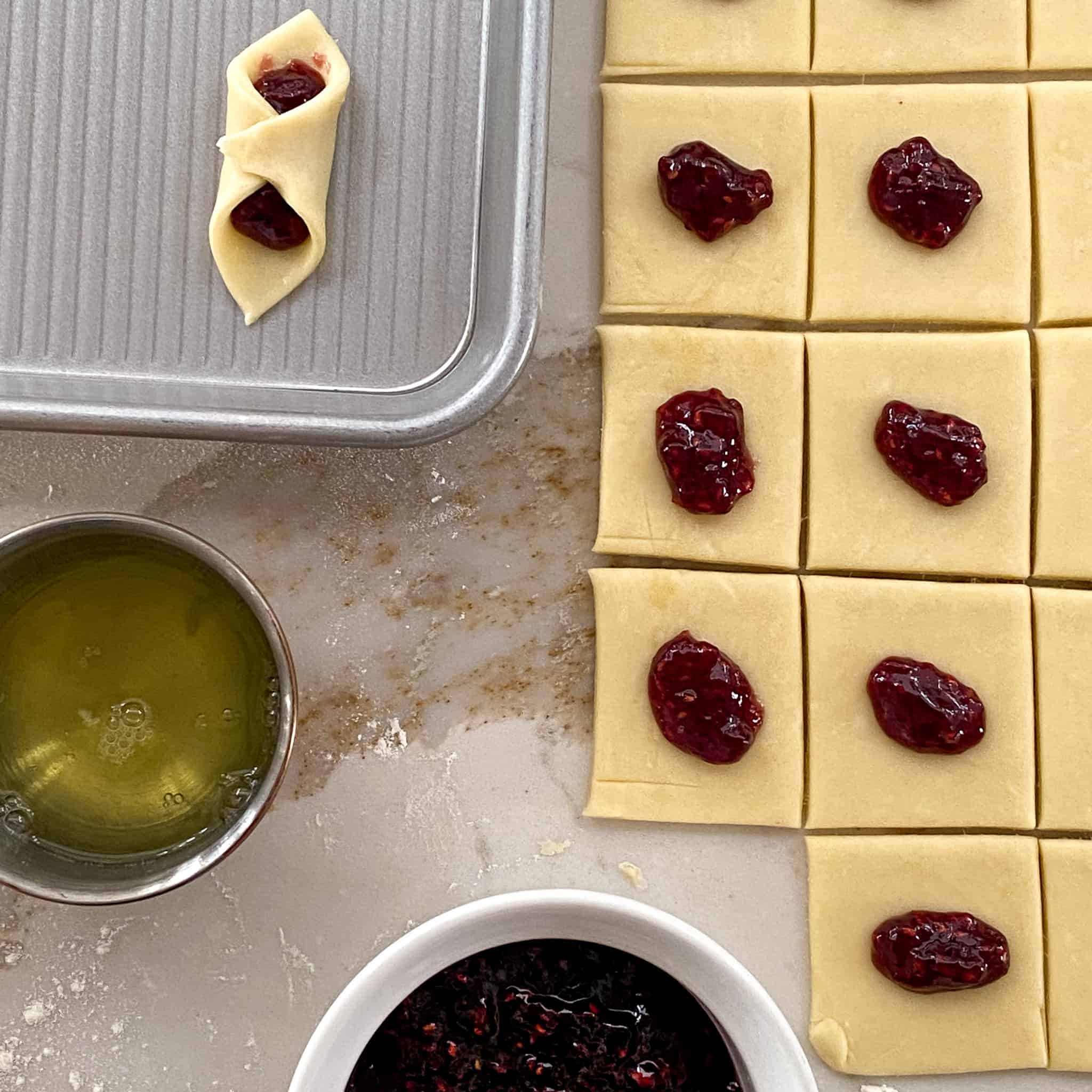 Jam spooned onto dough squares with a bowl of jam, egg white, and a folded kolaczki on a baking sheet.