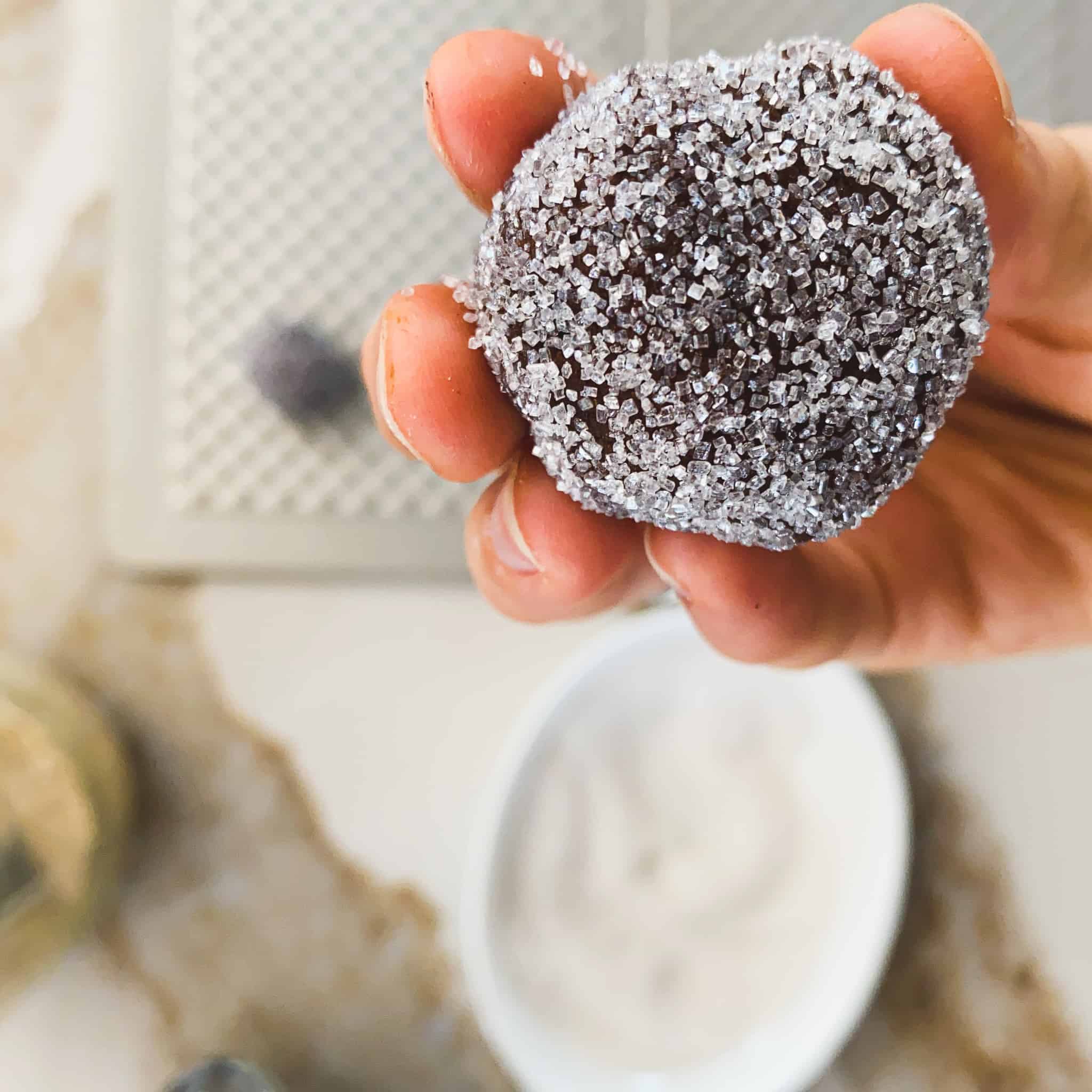 Raw cookie ball rolled in sugar and being held up over a cookie sheet with sugar in the background.