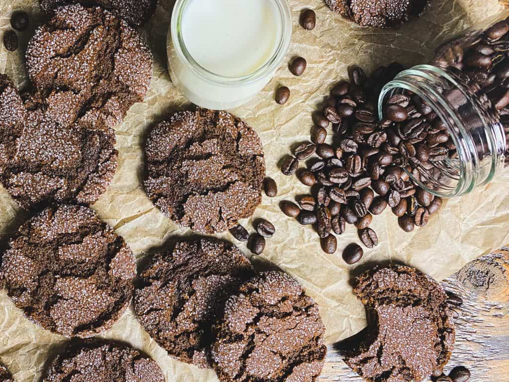 Dark brown espresso cookies with decorative sugar and large cracked tops. Cookies are on top of wood and parchment with coffee beans spilling out of a jar. A small jar of milk sits in the foreground.