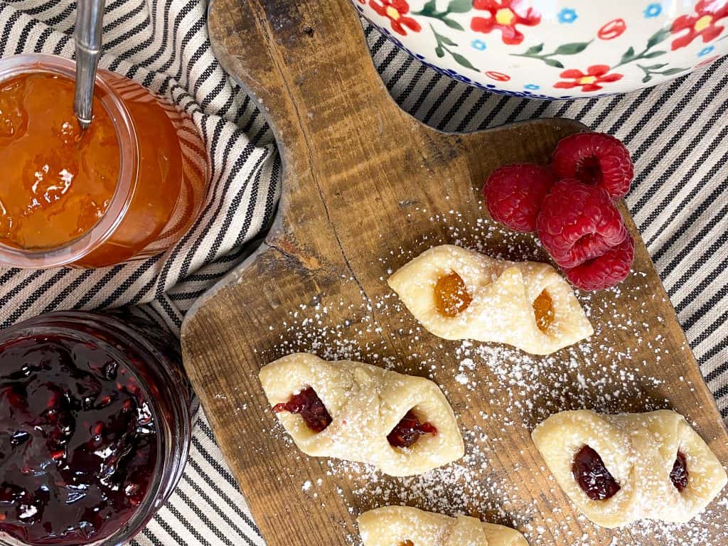 Baked Kolczki cookies on a wooden butcher block with a teapot, raspberries and raspberry jam in the foreground. A flowered polish teapot is peaking into the shot.