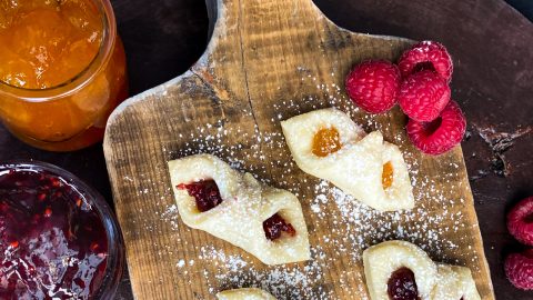 Icing sugar overtop of finished kolaczki cookies. The cookies are sitting on a rich wooden block with raspberries and apricot and raspberry jam on the side.