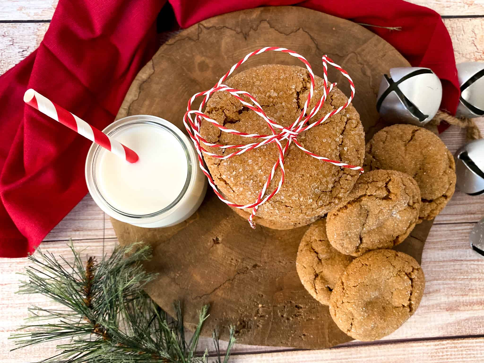 Old Fashioned gingersnap cookies on a large wooden slab. Crackled cookies are tied up with red and white string. There is a glass of milk, silver bells, and a sprig of spruce as well as a red linen napkin in the foreground.