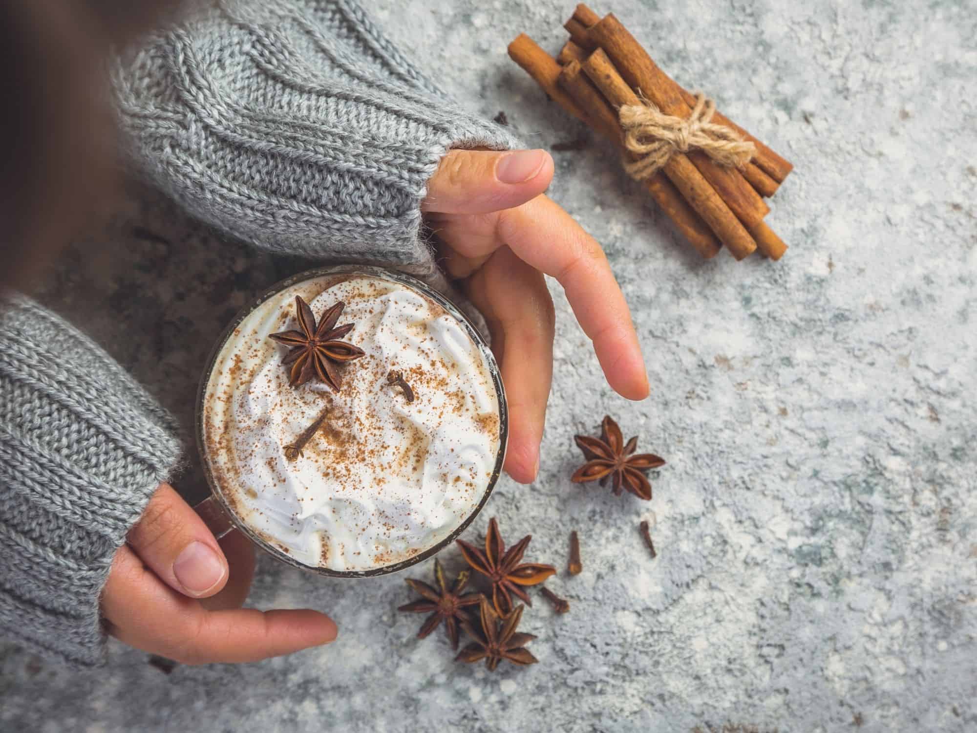 Hands holding a warm cup of chai tea latte with cinnamon sticks and star anise.