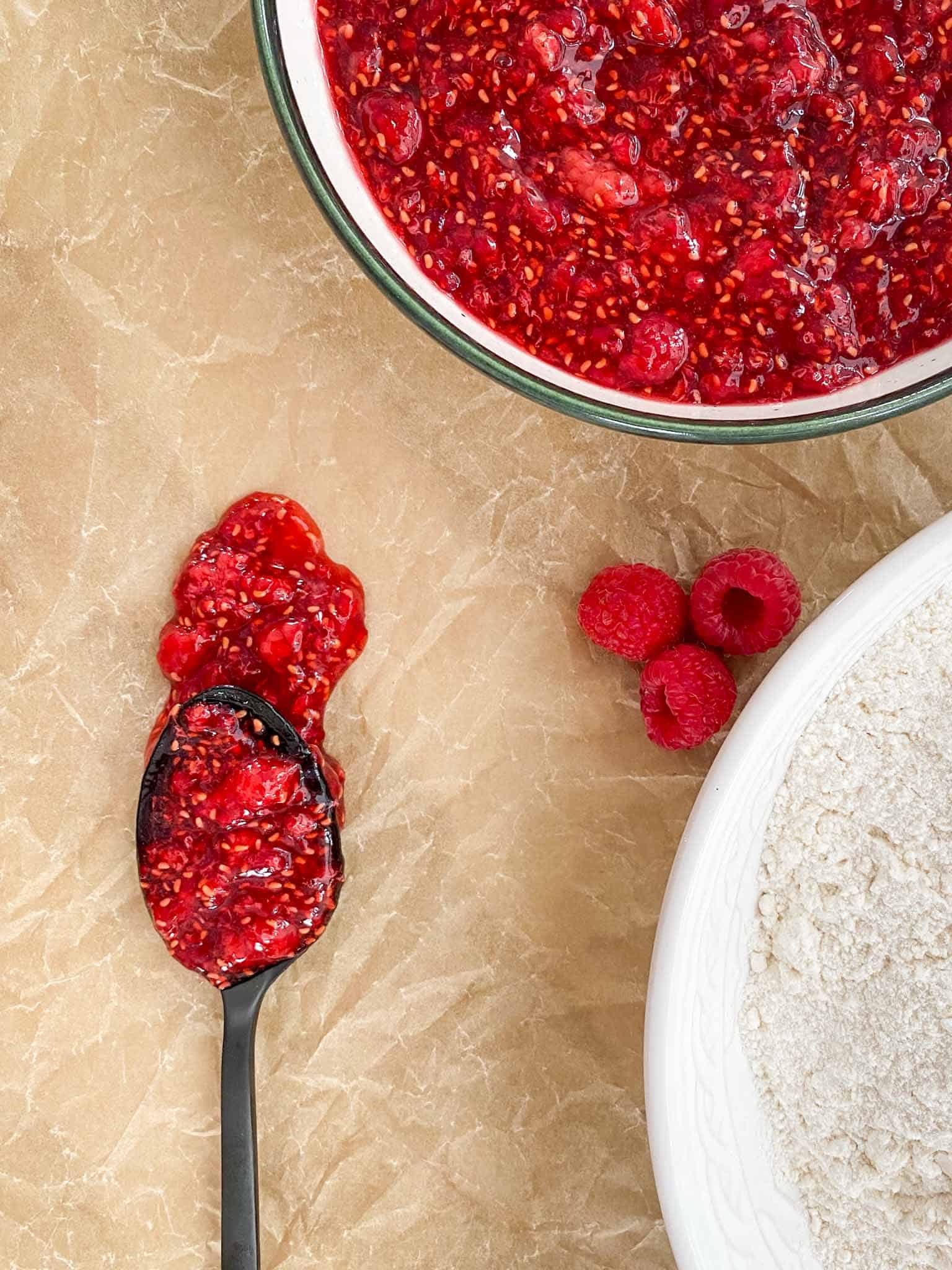 Red raspberry jam spilling off of a spoon in a large bowl.