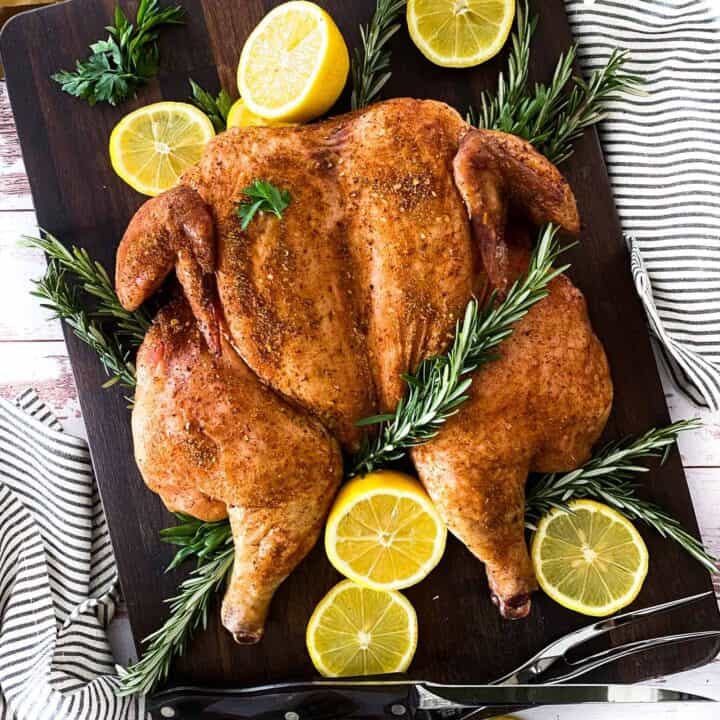 Smoked Spatchcock chicken on a large cutting board surrounded by rosemary, parsley, and lemons.