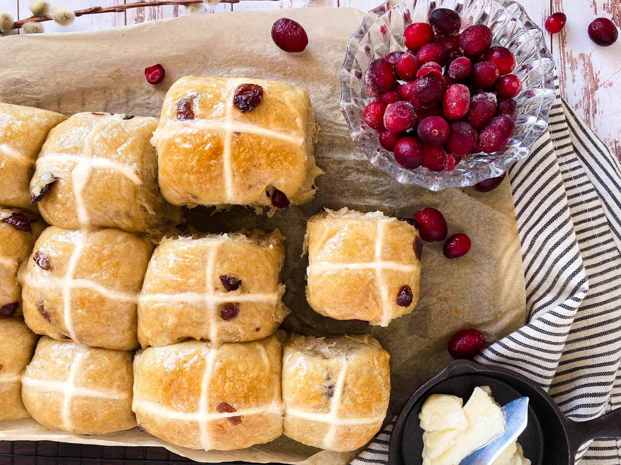 Overhead view of sourdough hot cross buns including cranberries in a decorative glass bowl and butter in a cast iron dish.