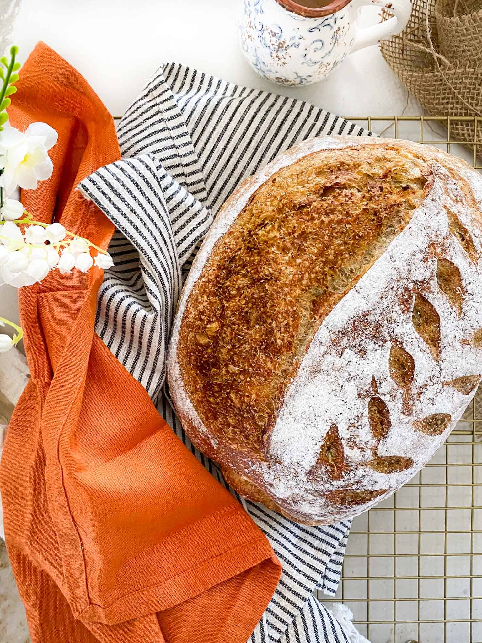 Wholegrain Sourdough Bread on a metal rack, surrounded by an orange and striped napkin and some flowers.