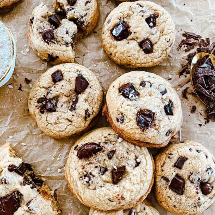 Chocolate chip cookies without butter piled on parchment paper and a golden cooling rack with chocolate and salt accenting.