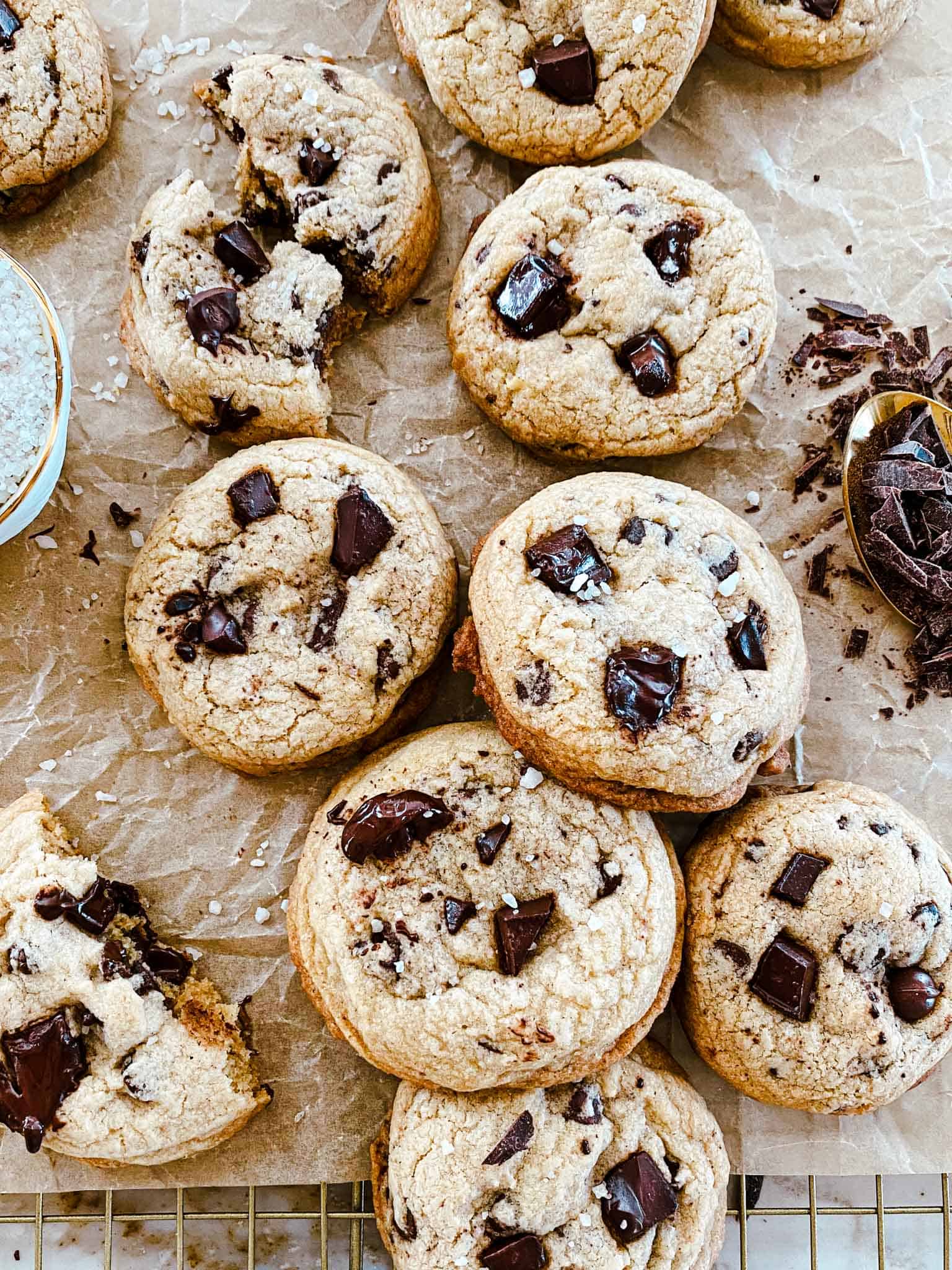 Chocolate chip cookies without butter piled on parchment paper and a golden cooling rack with chocolate and salt accenting.