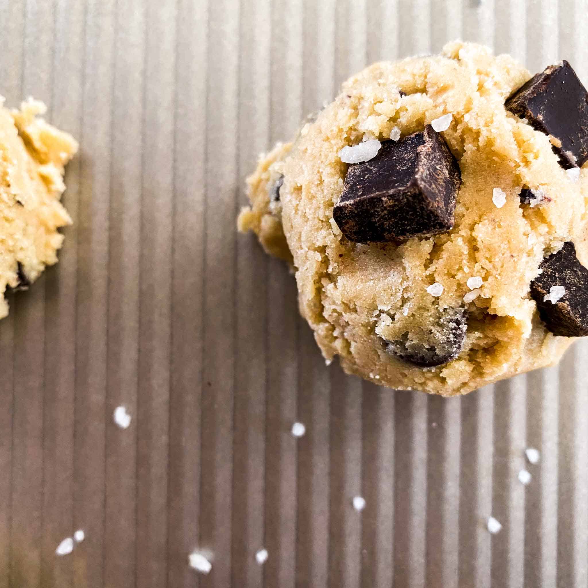 Two raw chocolate chip cookie dough balls on a baking sheet sprinkled with salt.