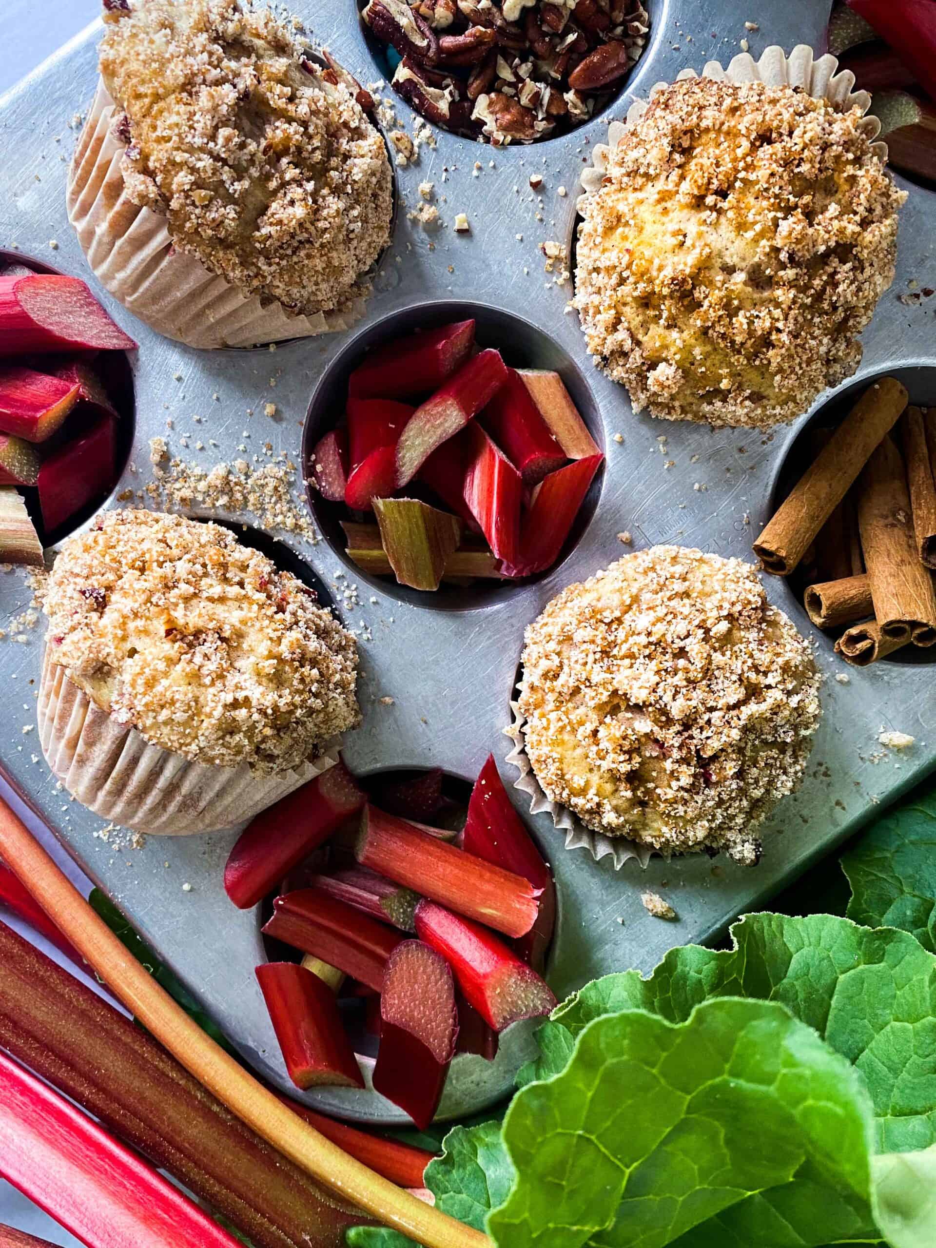 Rhubarb buttermilk muffins in a muffin tin with sliced rhubarb and rhubarb stalks with leaves behind. Cinnamon sticks and chopped pecan accompany the rhubarb muffins.