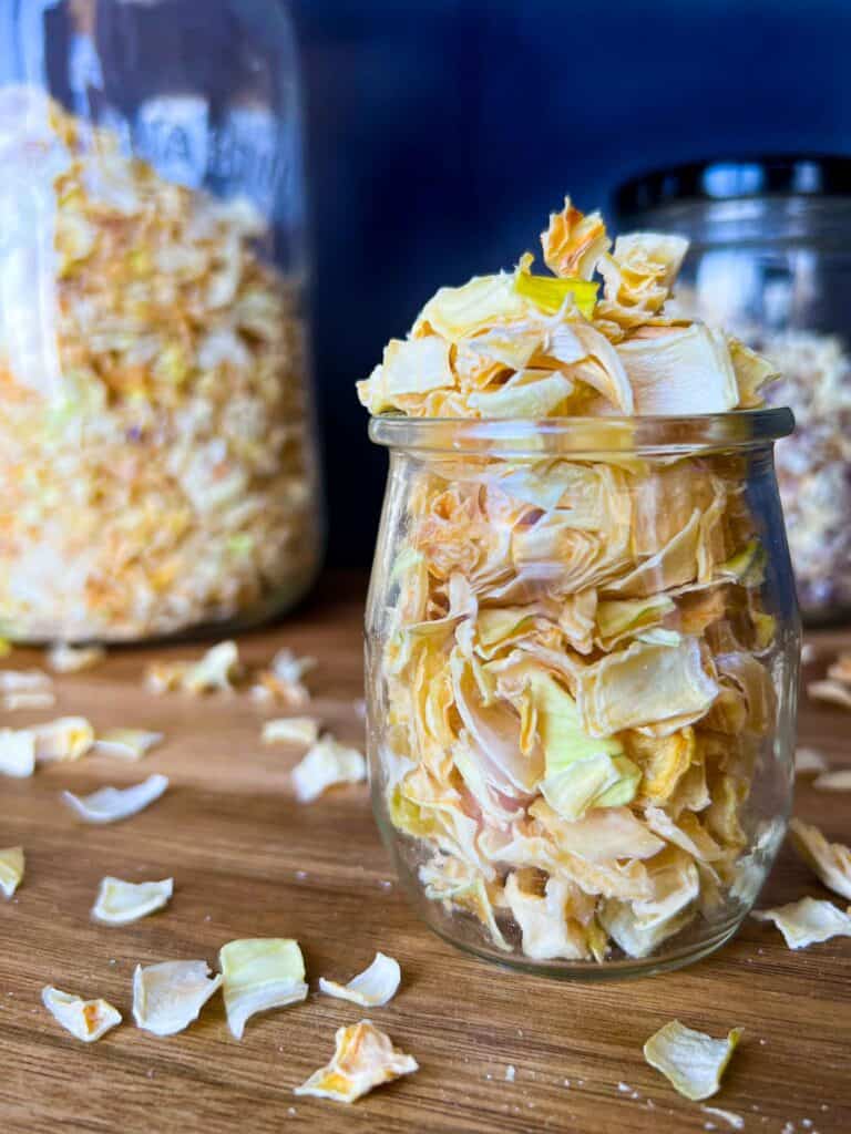 Front view of dehydrated onions in a glass jar on a wooden cutting board with dried onions scattered around and tall jars in the foreground.