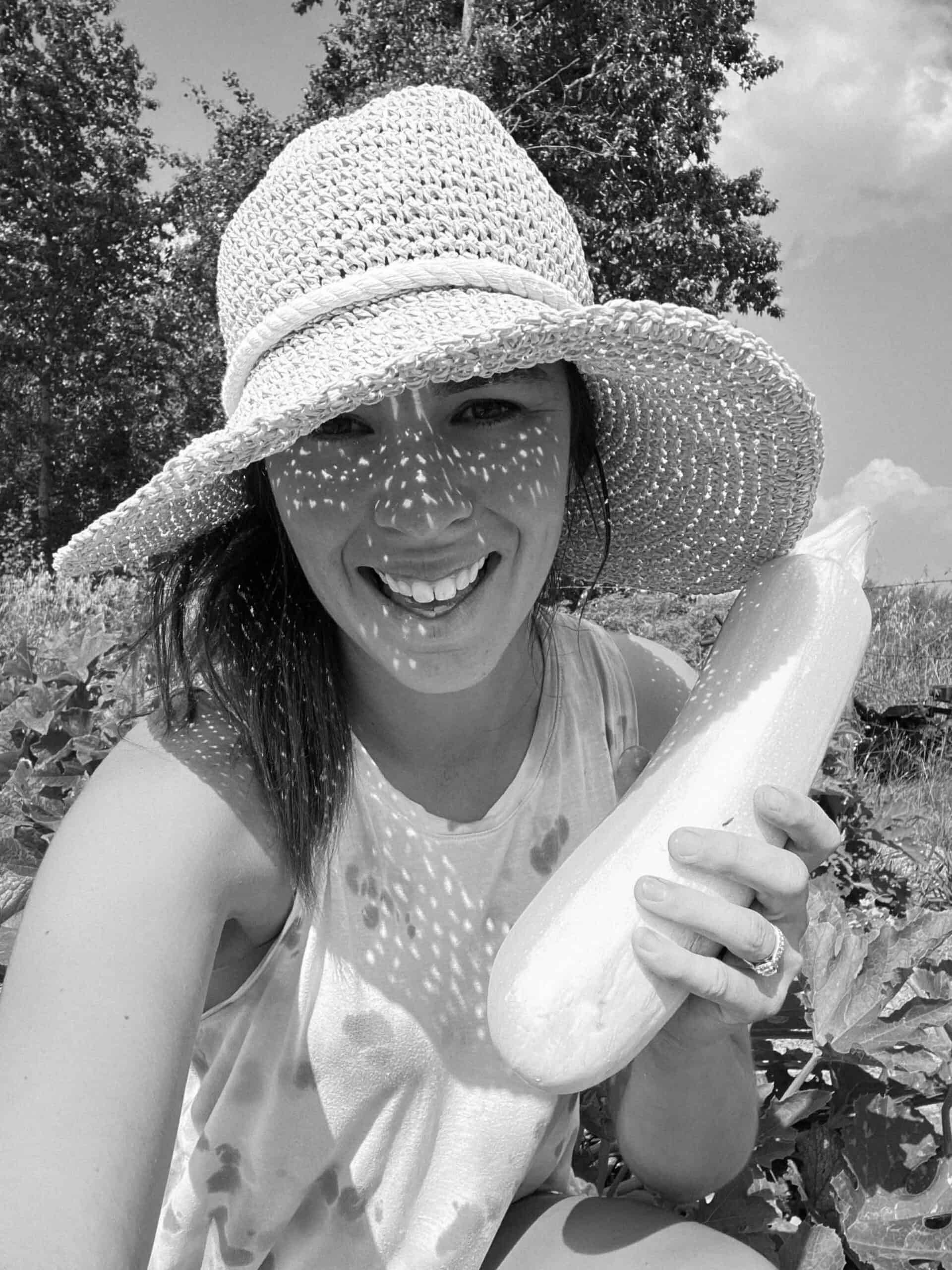 Kate (content creator) holding a zucchini in the garden with a large floppy hat.