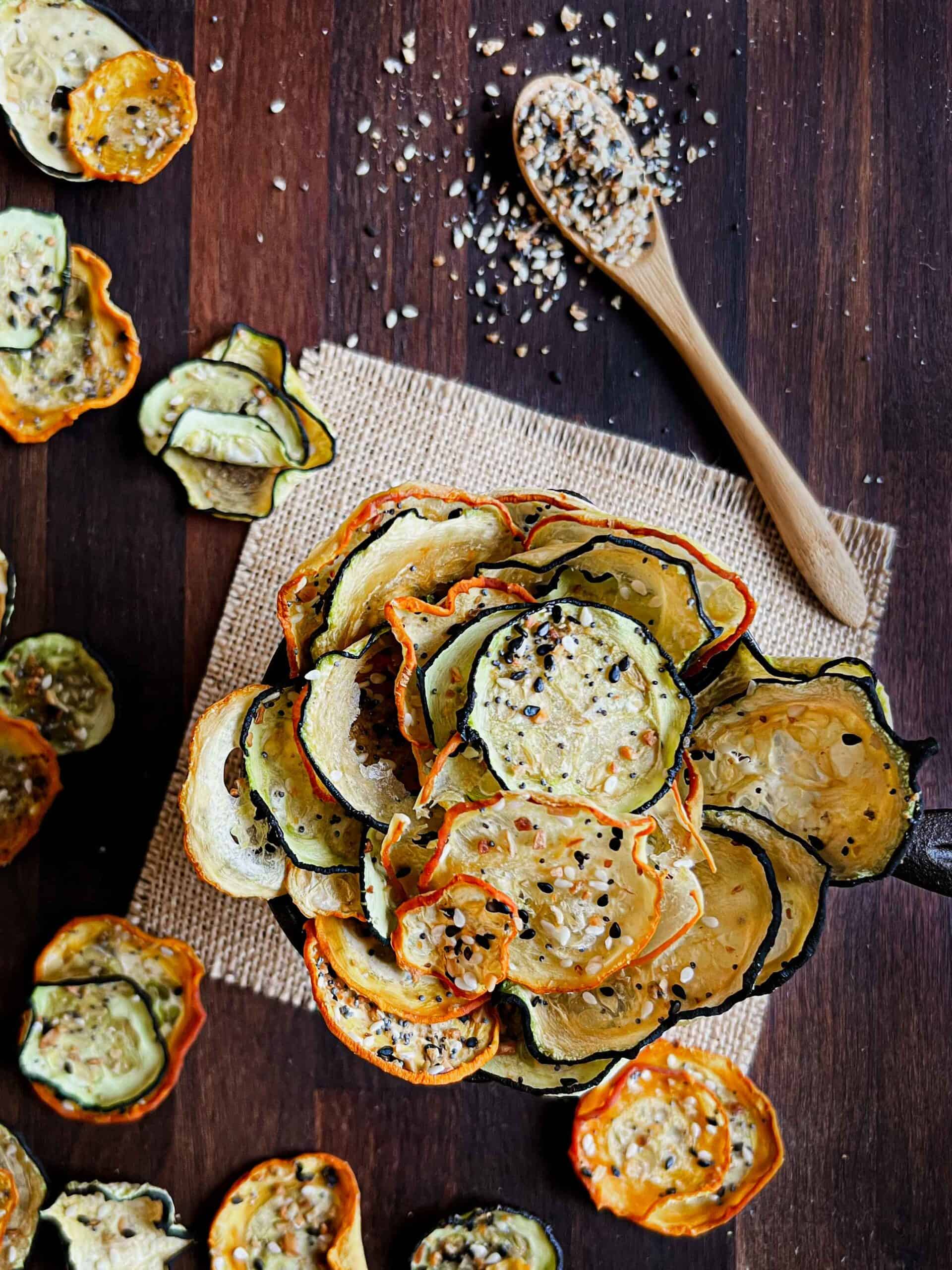 Everything bagel spice zucchini chips in a small cast iron pan sitting on burlap and a wooden cutting board with chips all around.