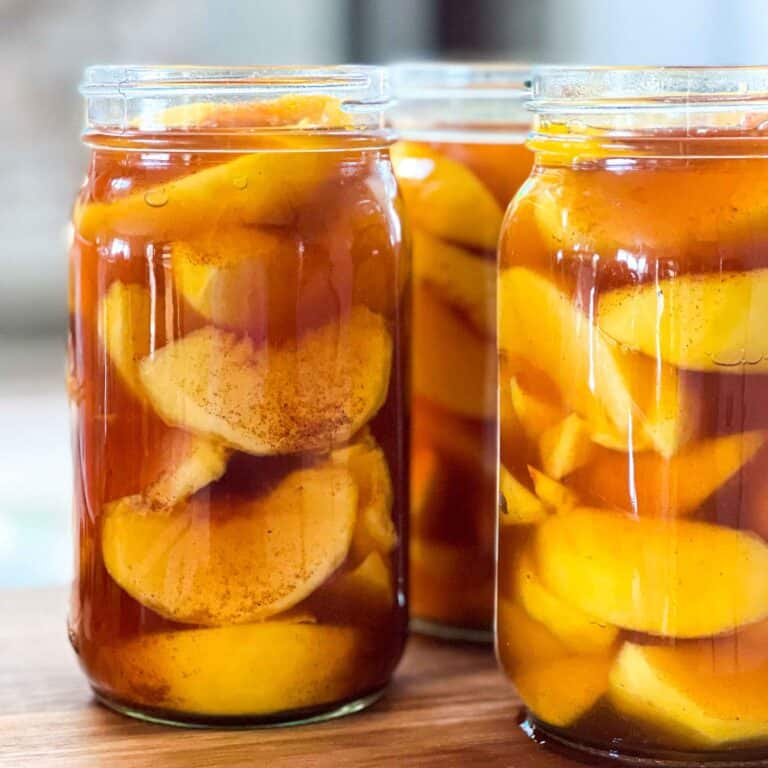 Southern-style canned peaches with brown sugar and spices in three jars on a wooden cutting board.
