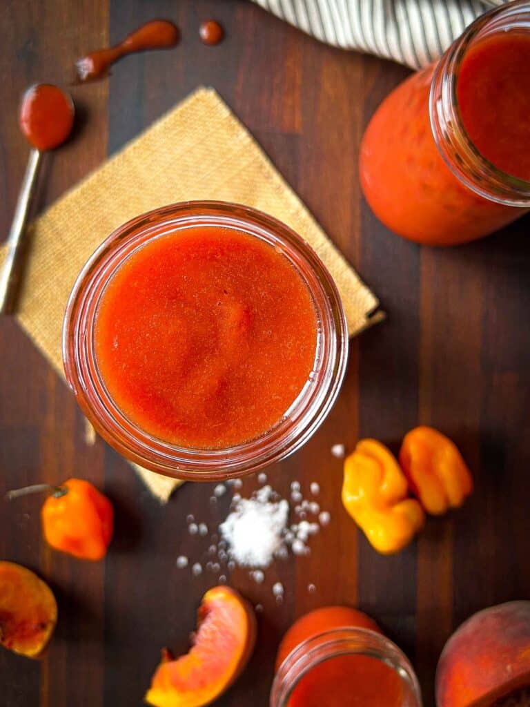 Smoked peach habanero hot sauce in jars with salt, peppers, peaches, and spilled sauce on a wooden cutting board.