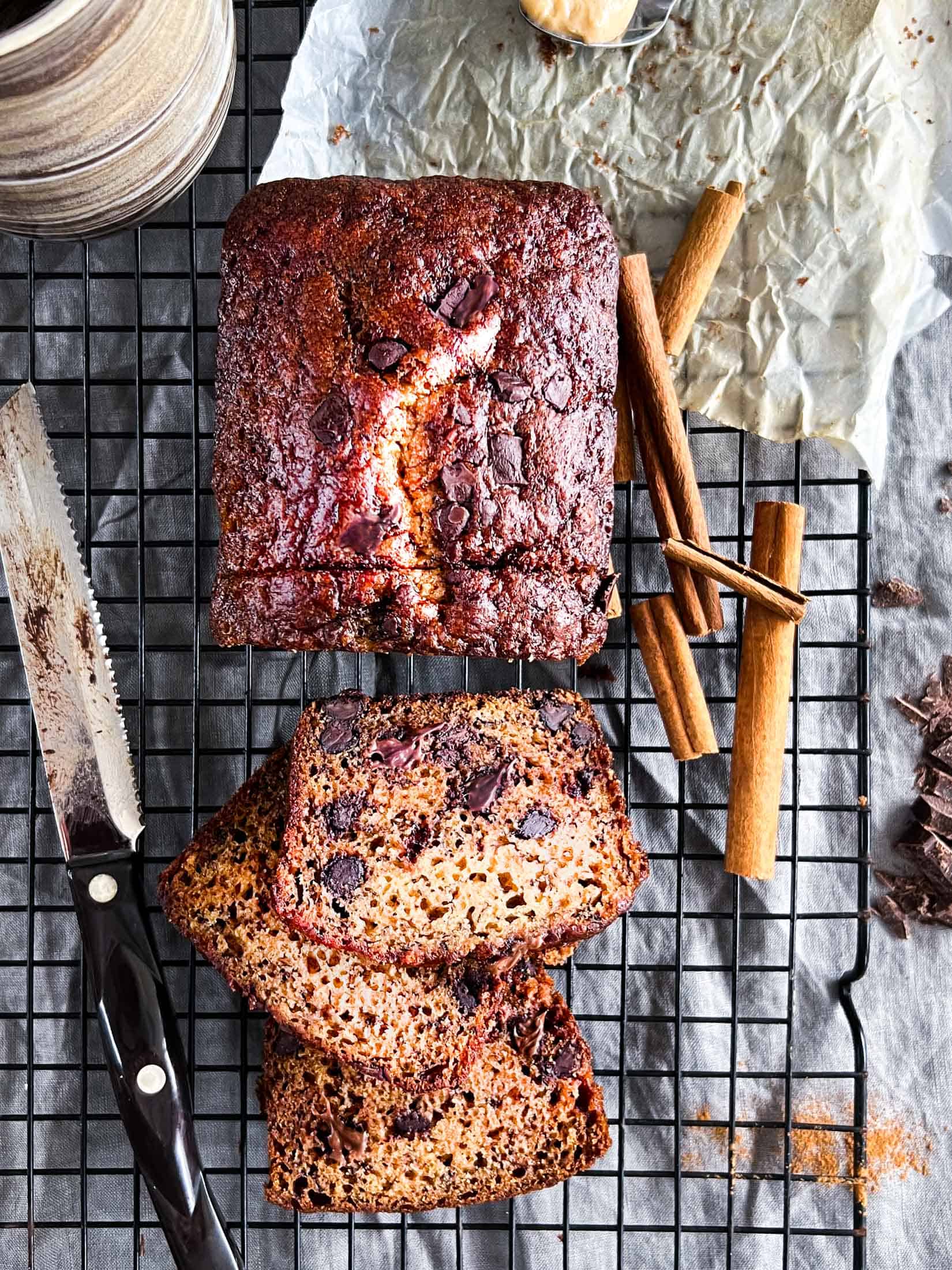 Banana bread without butter baked and sitting on a piece of parchment and cooling rack with cinnamon sticks, a chocolate smeared knife, and cinnamon sprinkled in the background.
