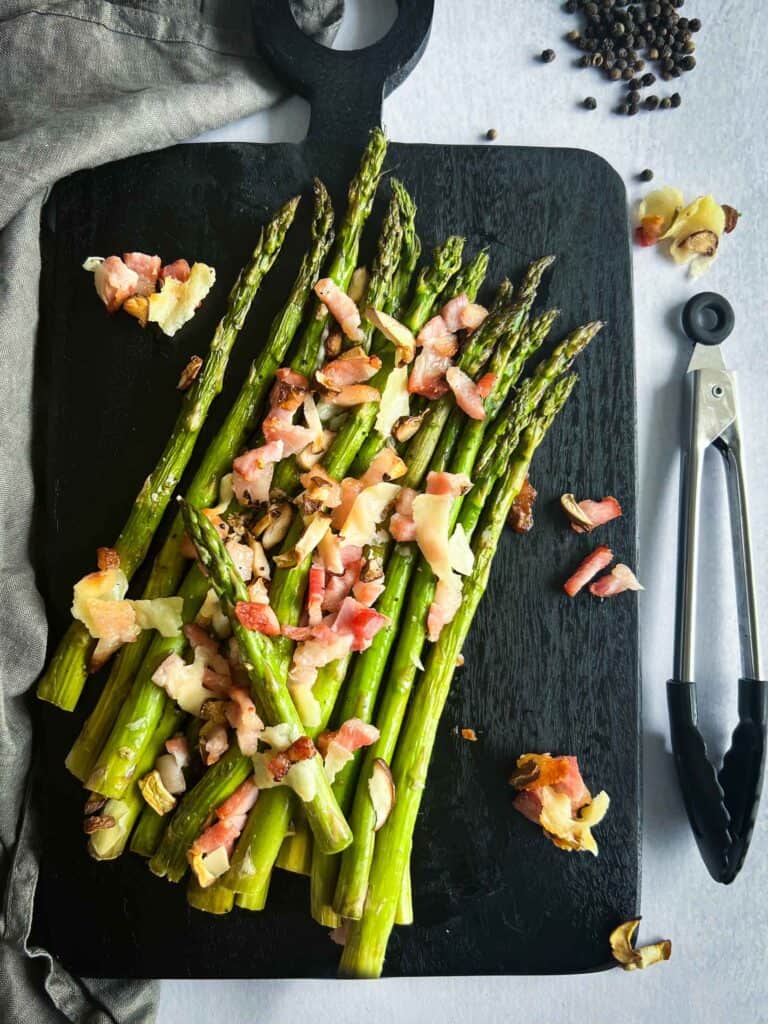 Traeger asparagus after being smoked, sprinkled with bacon, parmesan, mushrooms. Sitting on a black cutting board.