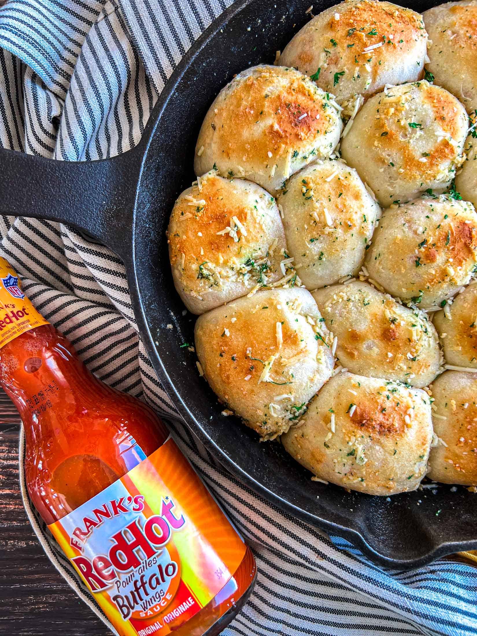 Fully cooked buffalo chicken bombs in a cast iron pan with browned tops and parmesan and parsley sprinkled overtop and a bottle of Frank's Red Hot Buffalo Wing Sauce nestled beside.