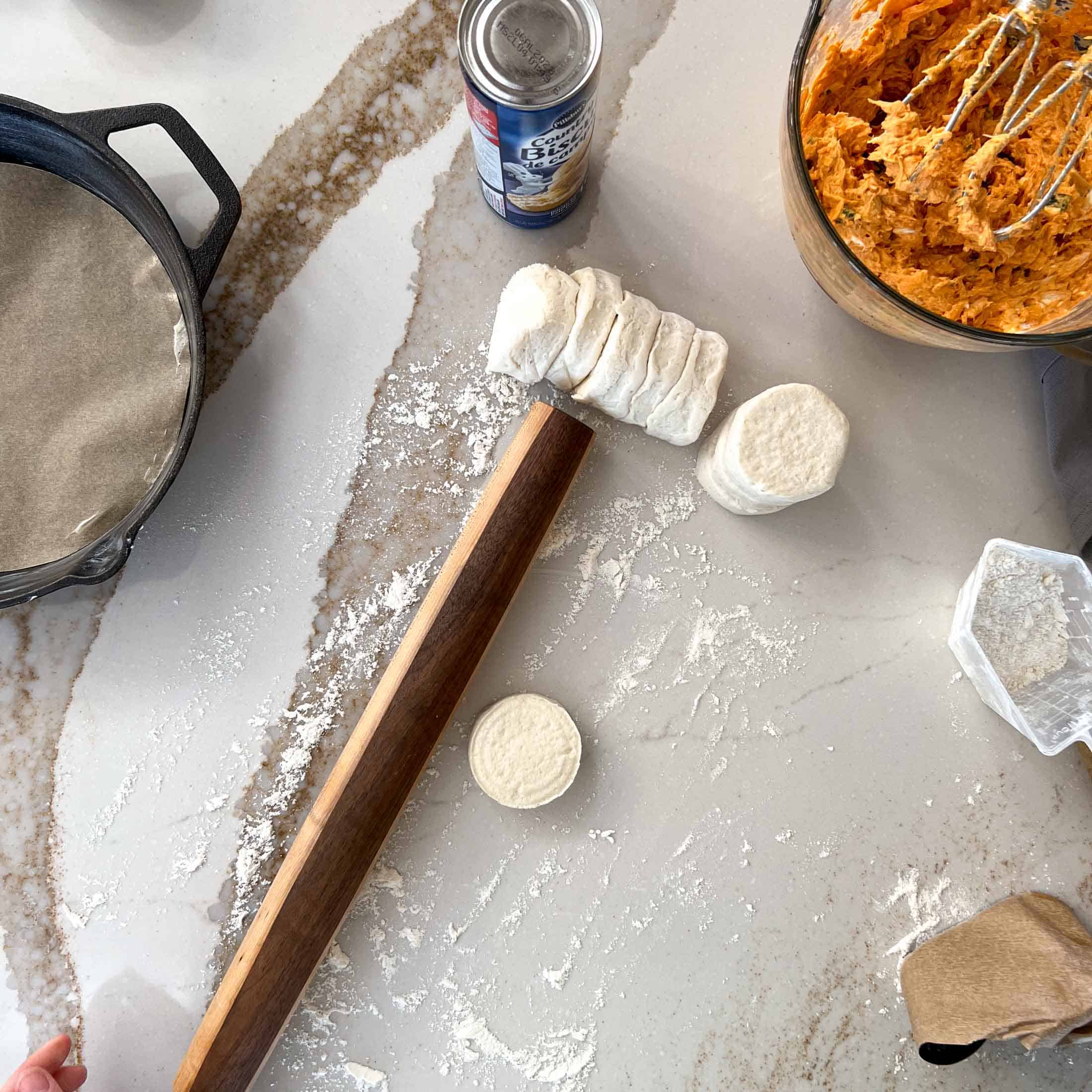 Refrigerator biscuits sitting on a floured countertop with a rolling pin and other ingredients.