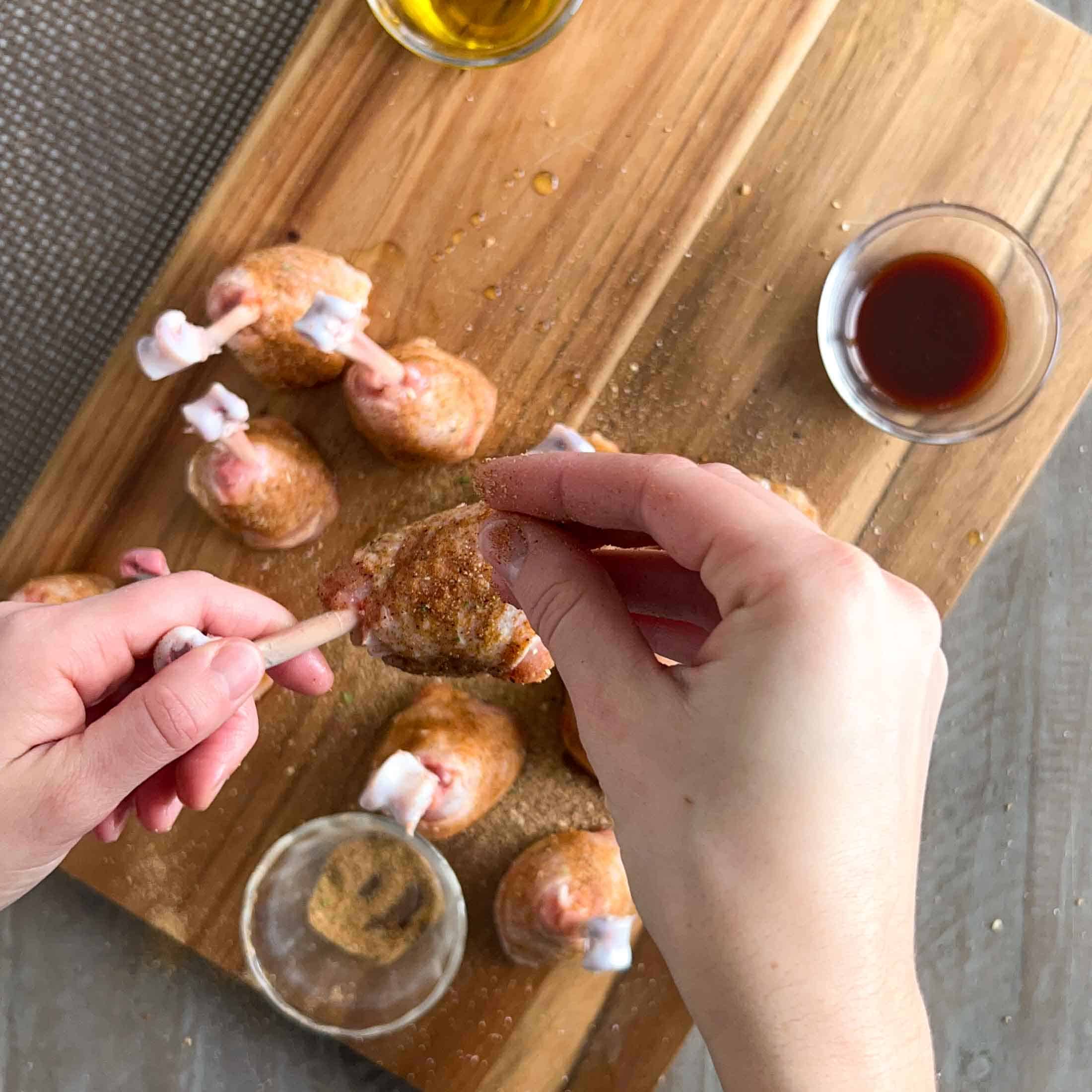 A hand sprinkling rub onto a prepared chicken lollipop before grilling.