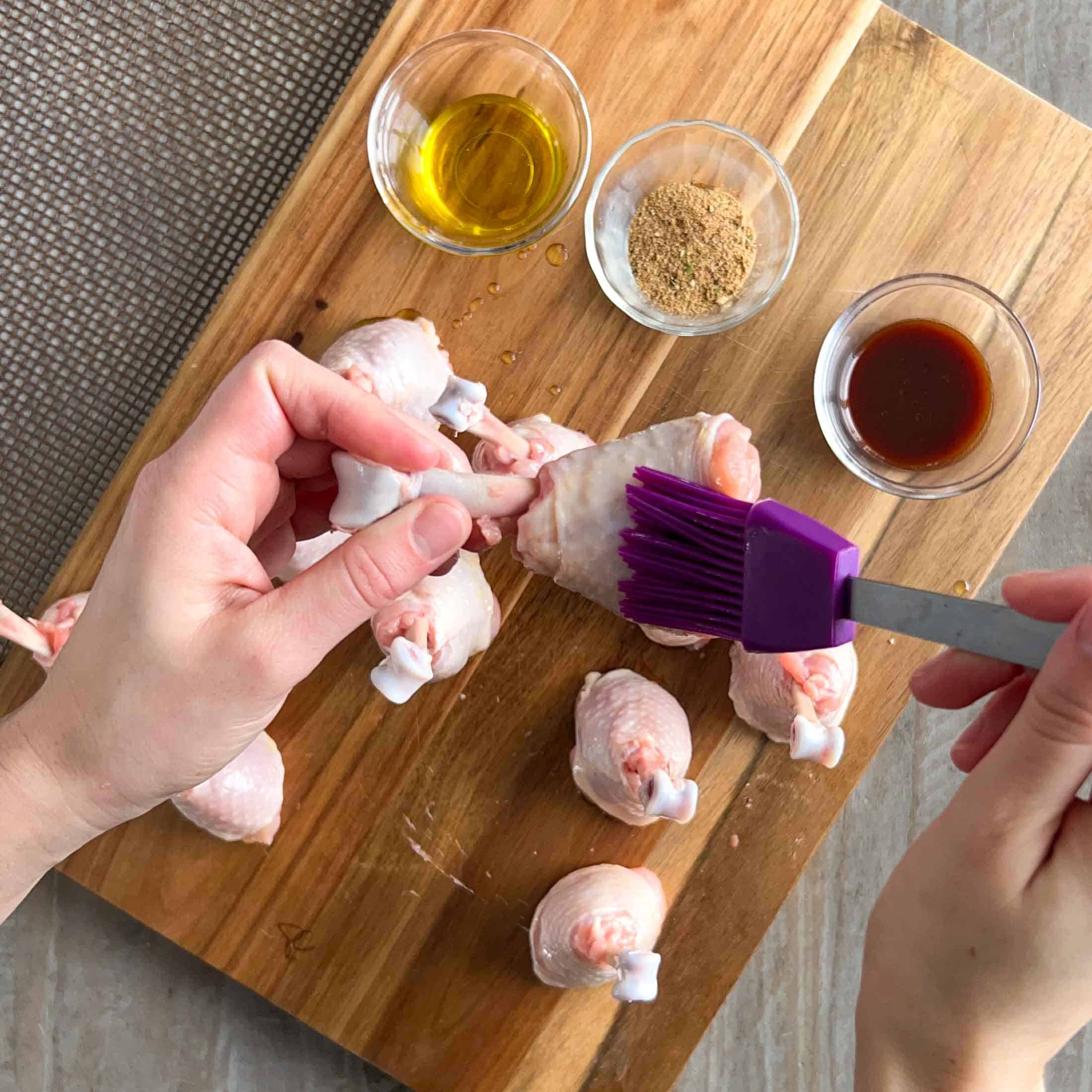 A purple basting brush, brushing on olive oil to a raw chicken lollipop.