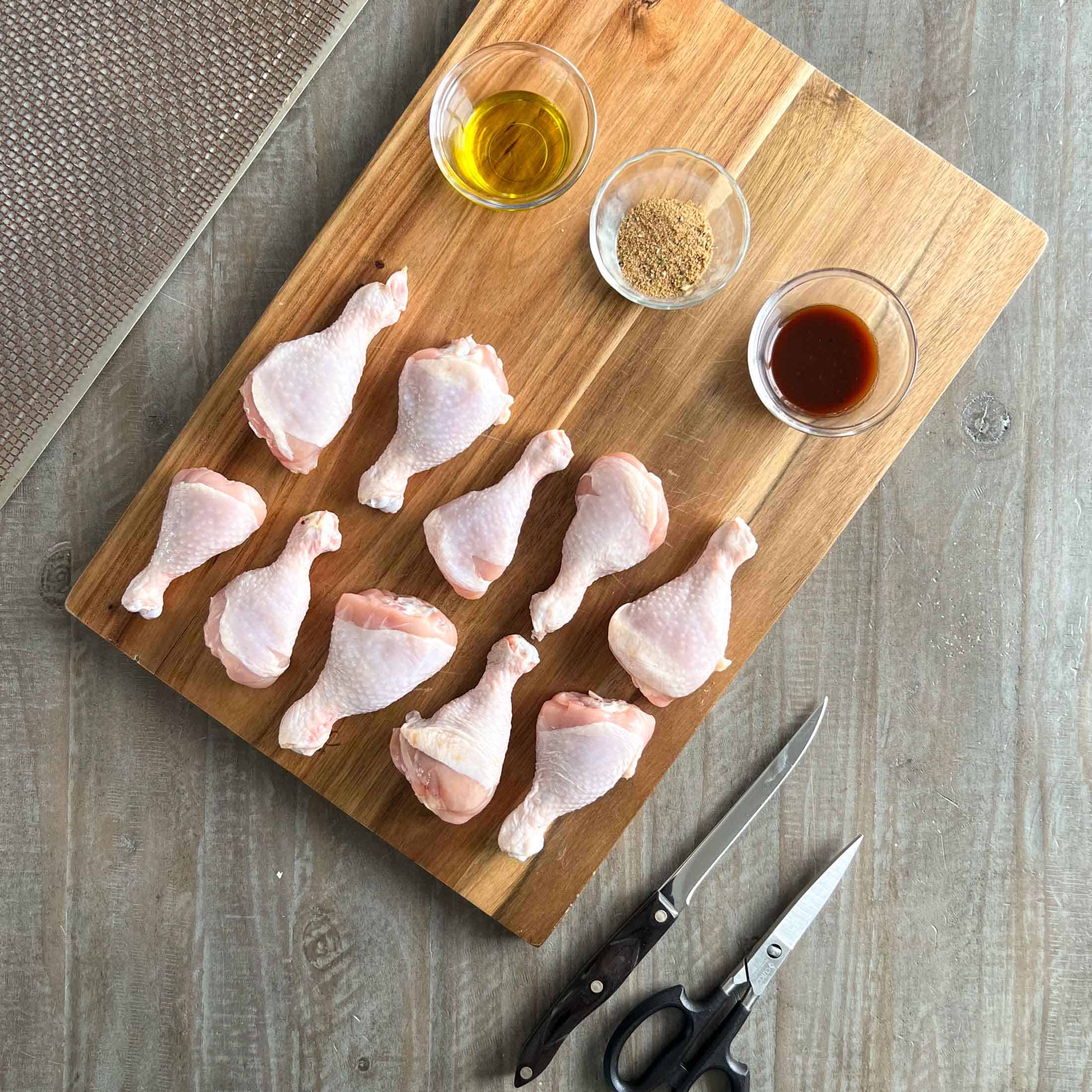 Ten drumsticks, olive oil, chicken rub, and barbecue sauce on a wooden board.