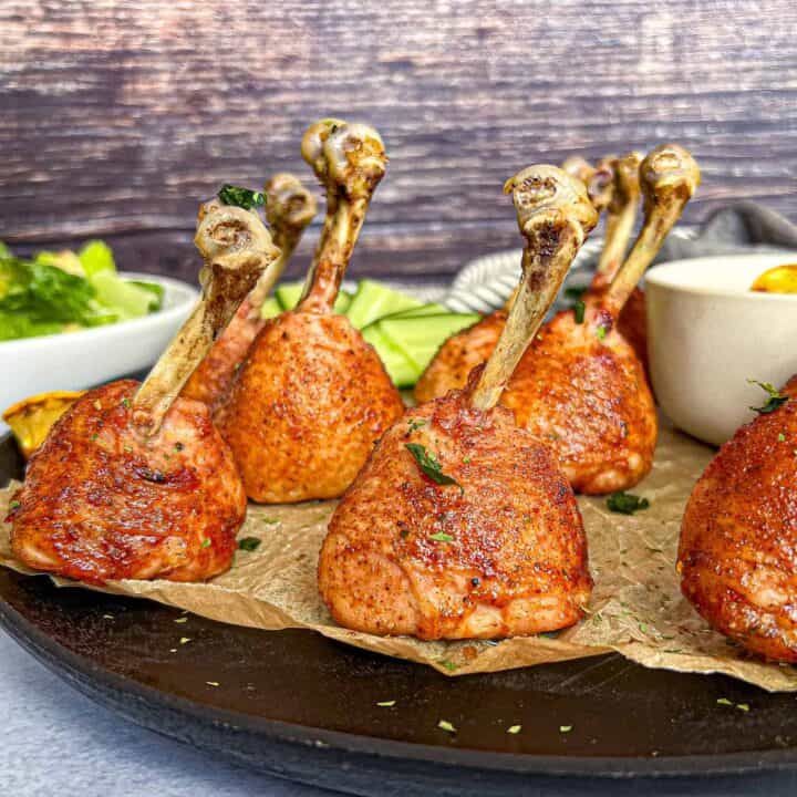 Crispy grilled chicken lollipop drumsticks on a black plate with brown parchment crinkled underneath.