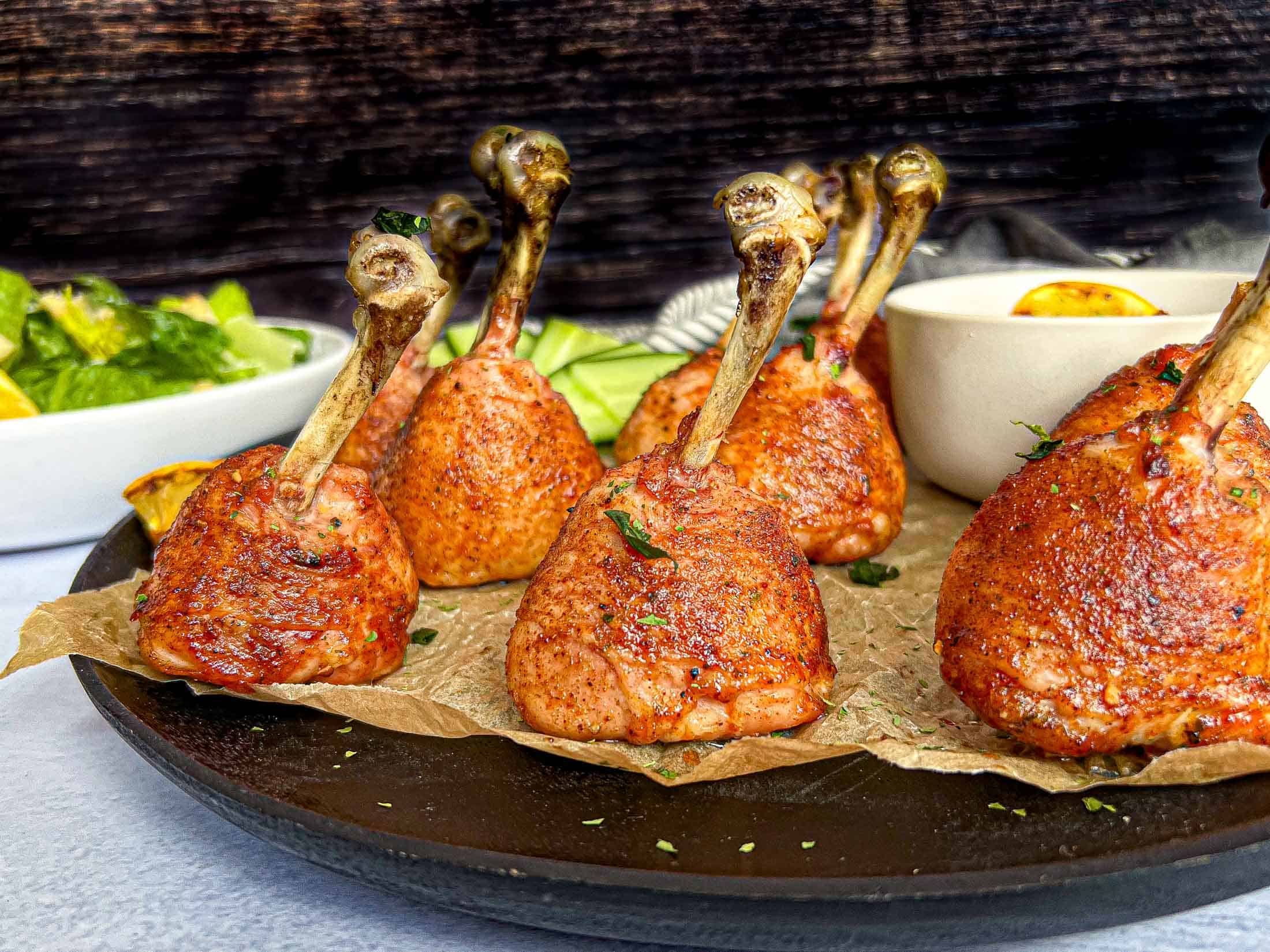 Crispy grilled chicken lollipop drumsticks on a black plate with brown parchment crinkled underneath.