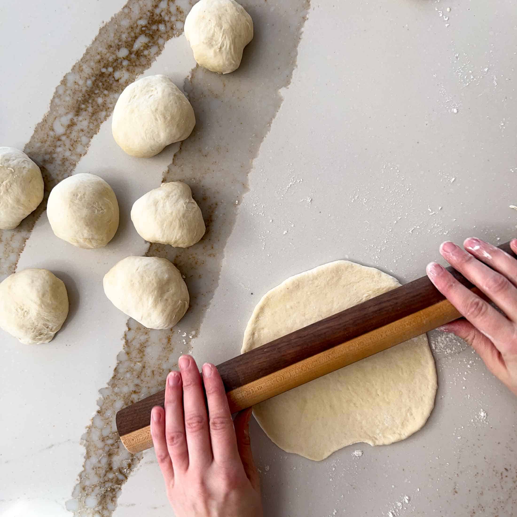 A wooden rolling pin rolling out a piece of naan bread with prepared dough balls beside.