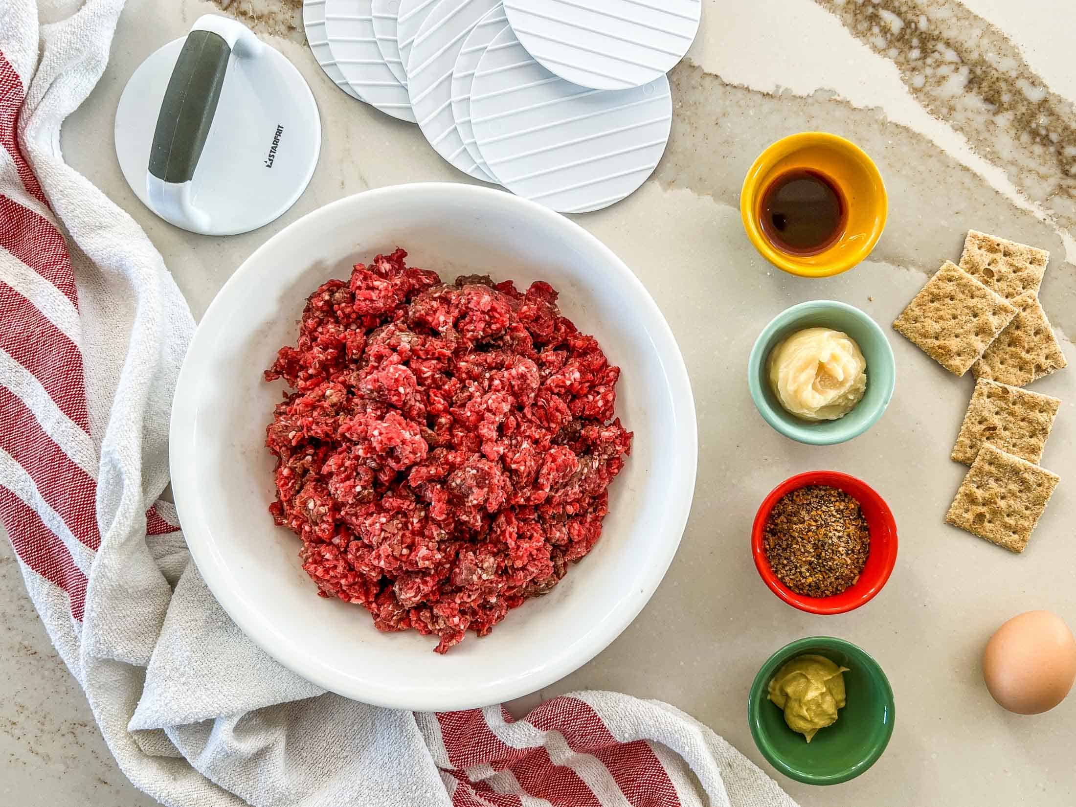 Key ingredients for venison burgers including ground deer meat, lard, steak spice, dijon mustard, Worcestershire sauce, crackers, and an egg.