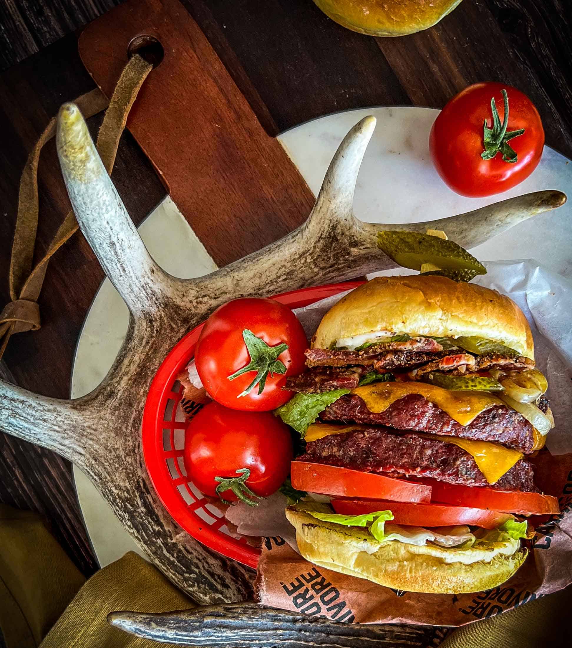 Deer burgers stacked together with cheese melted on top, sandwiched in between two buns and a lot of burger toppings.