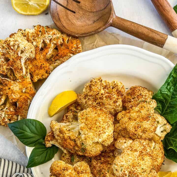 Smoked cauliflower steaks and cauliflower florets with large wooden utensils and lemon wedges.