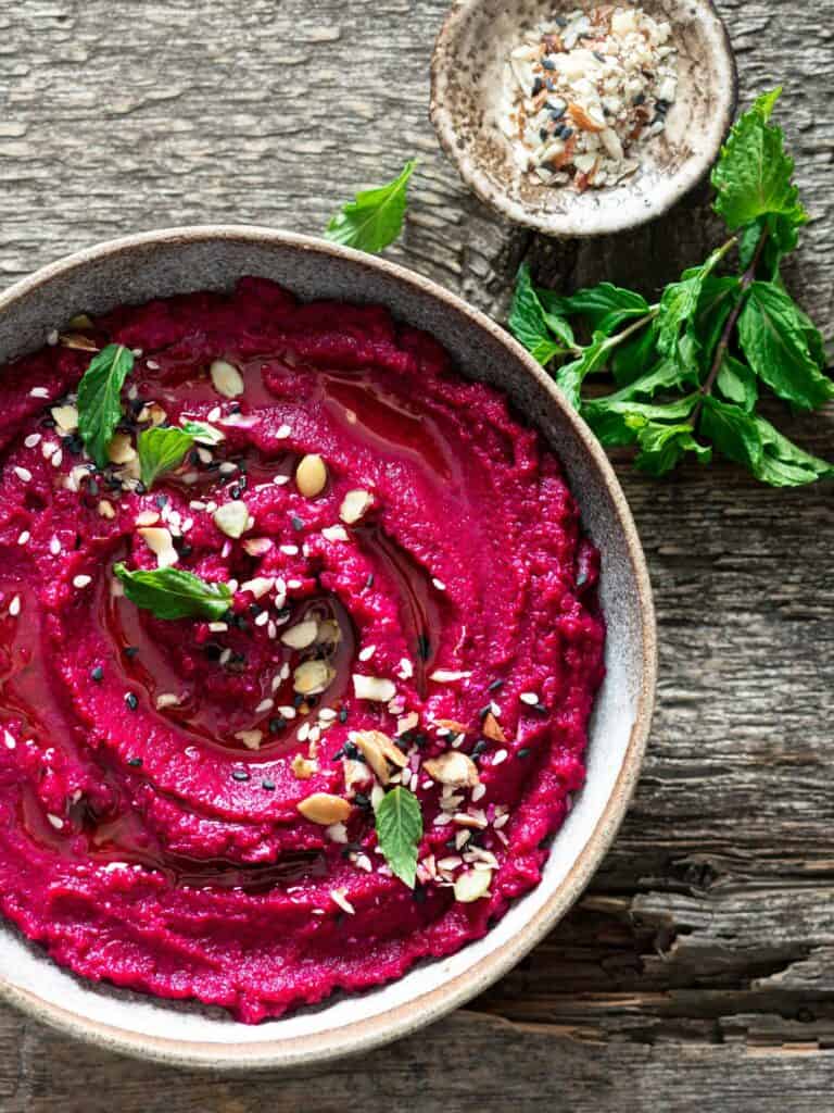 Smoked beet hummus in a stone bowl with herbs and seeds as garnish.