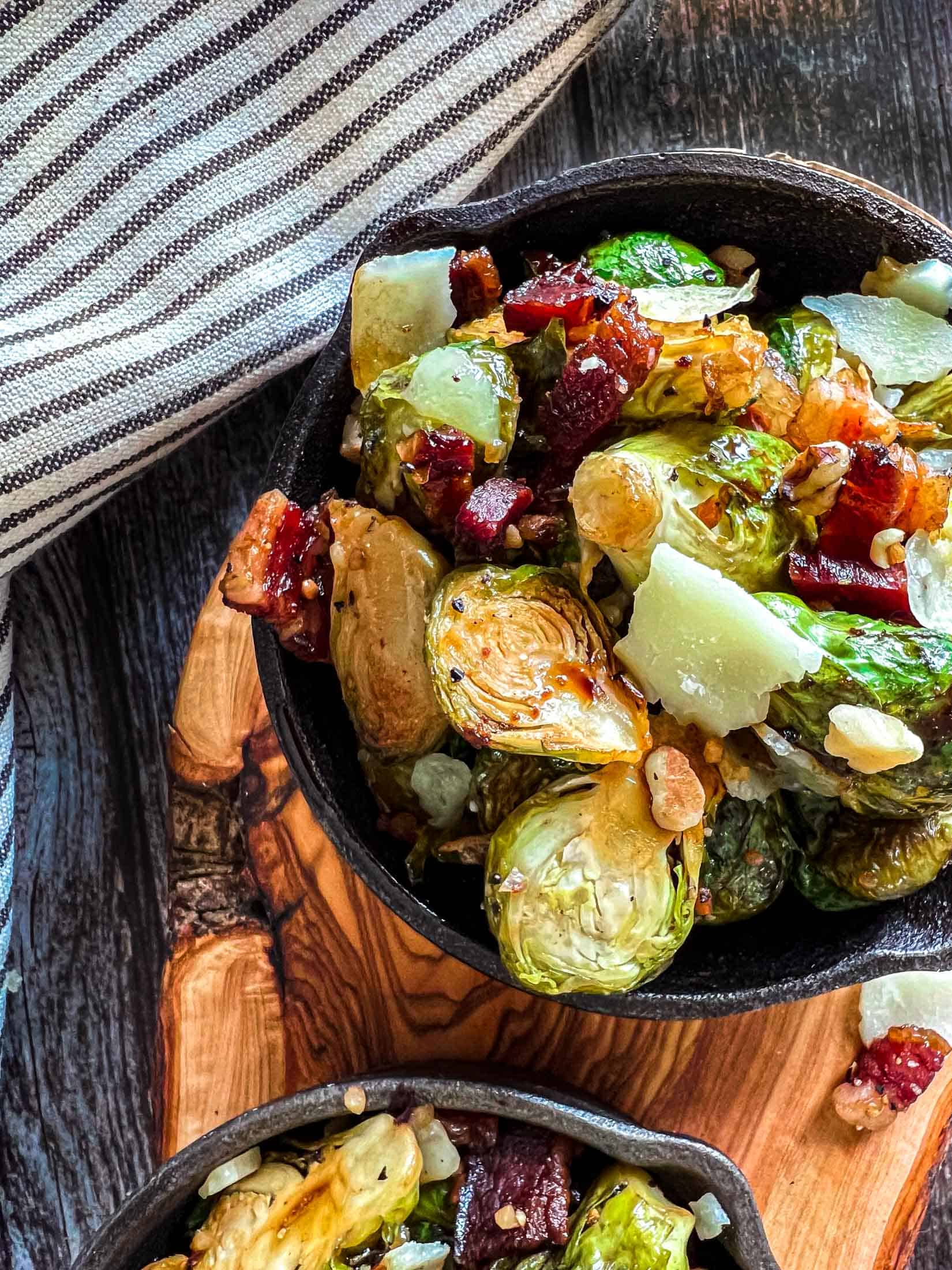 Smoked brussel sprouts with bacon, onions, and glaze in mini cast iron skillets on a wooden cutting board.