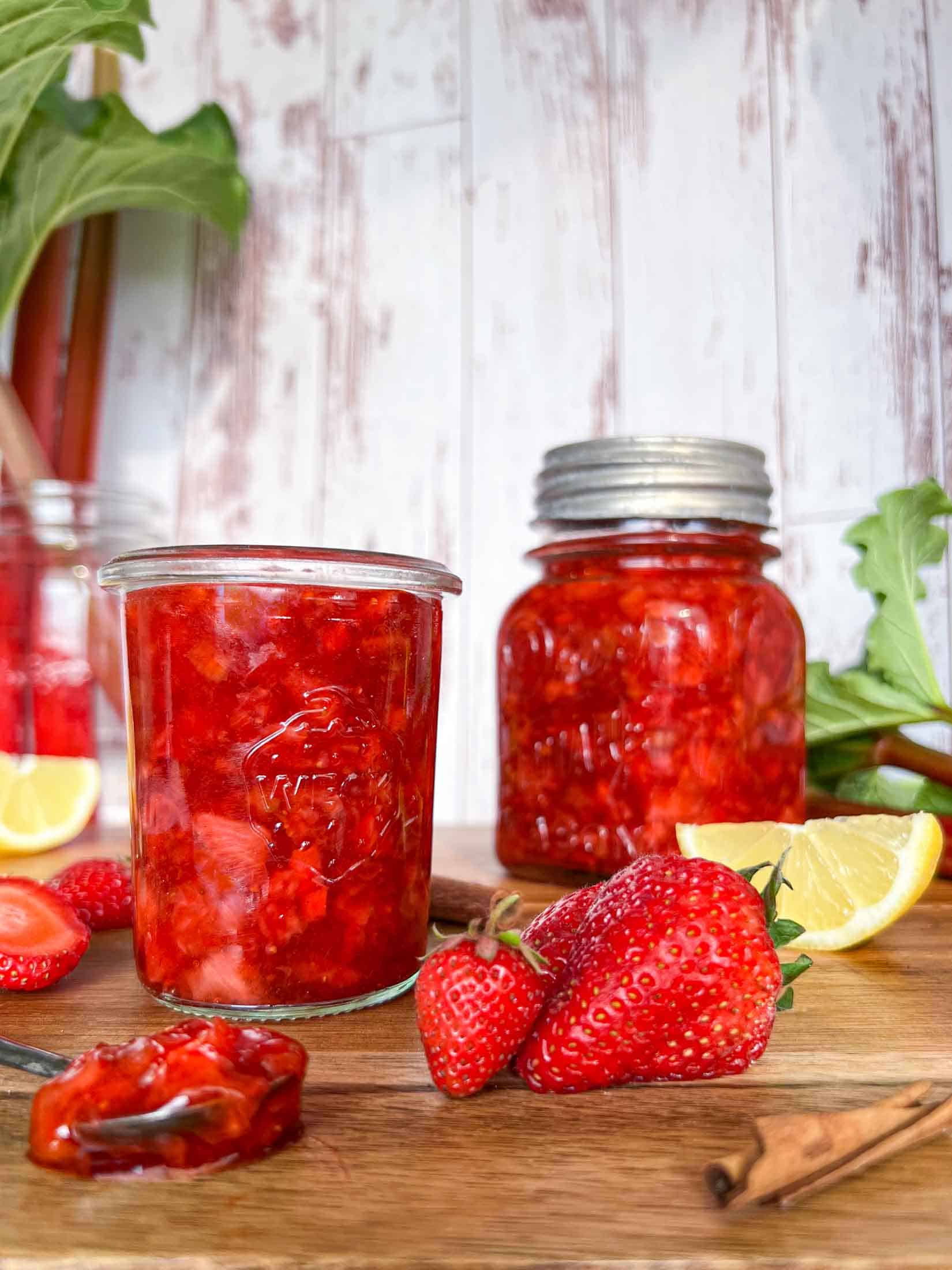 Strawberry rhubarb freezer jam in a small jar with another jar in the background. Sliced lemons and strawberries surround the jars on a cutting board with rhubarb in the background.