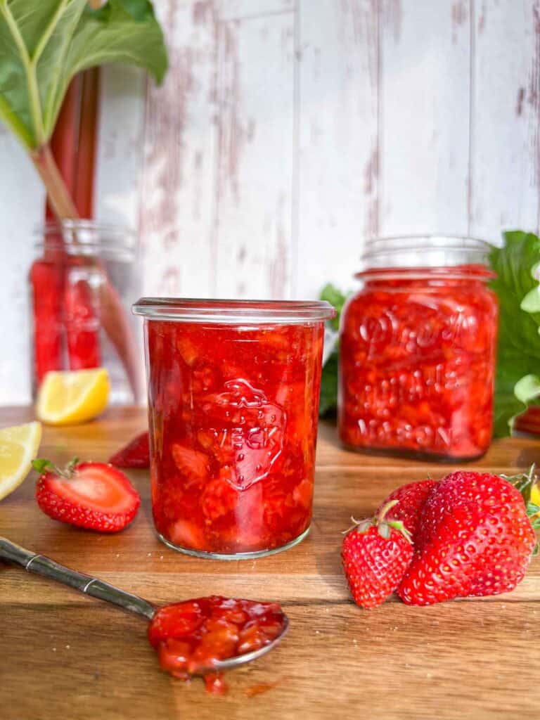 Strawberry rhubarb freezer jam in a small jar with another jar in the background. Sliced lemons and strawberries surround the jars on a cutting board with rhubarb in the background.