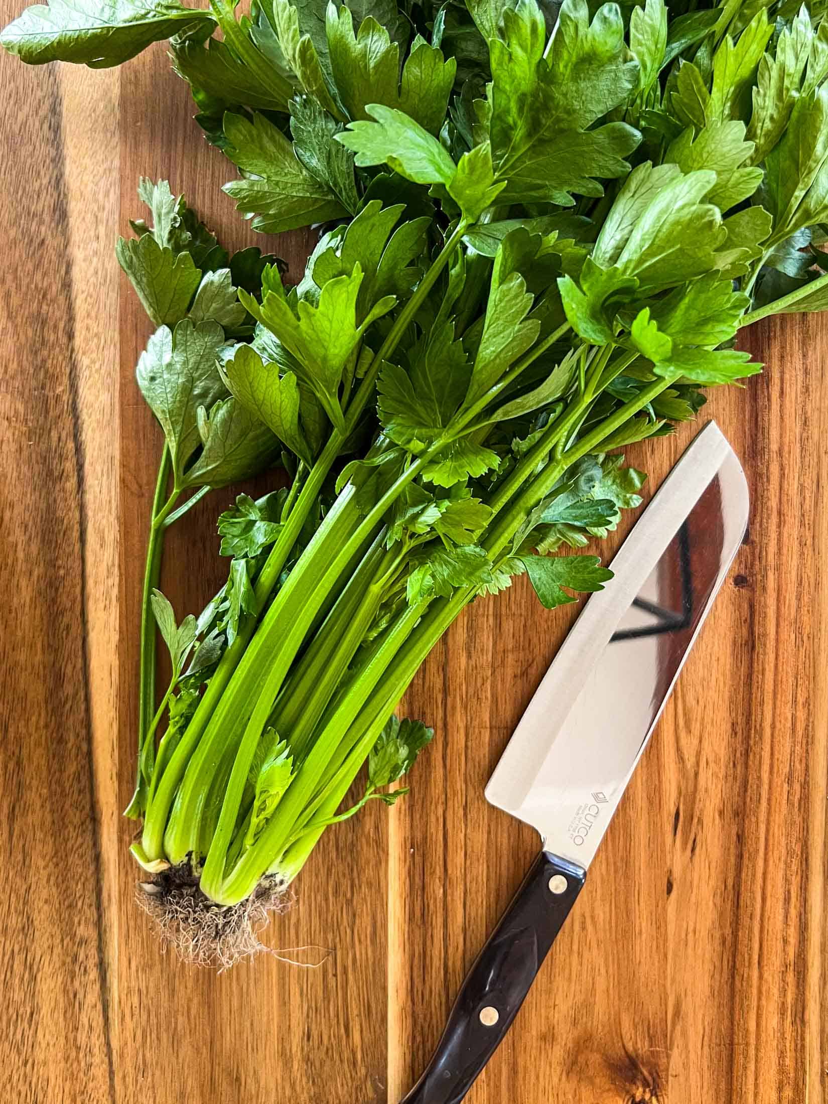 A large bunch of celery on a wooden cutting board with a chef's knife beside.
