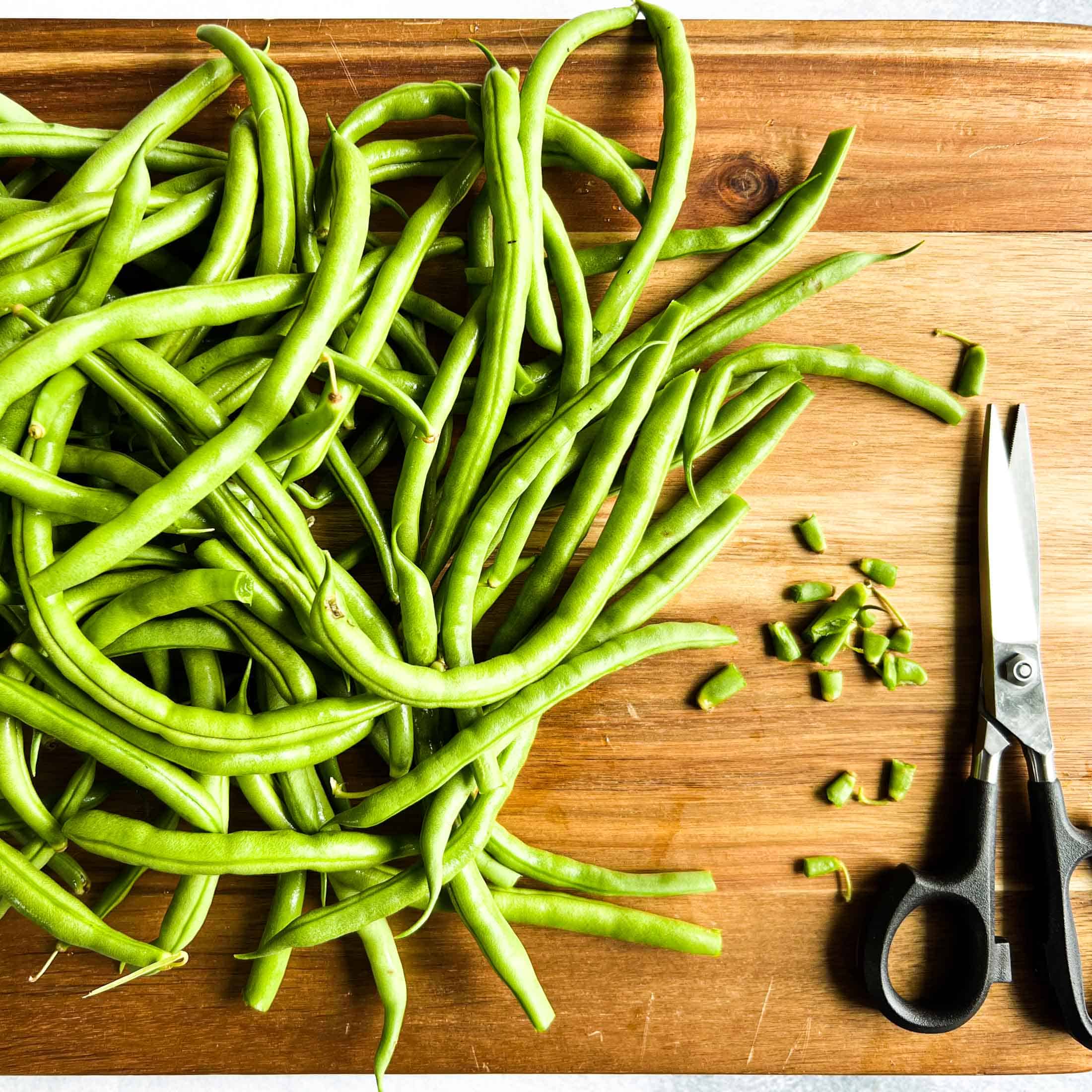 Green beans on a wooden cutting board with kitchen shears beside and little pieces of cut tops.