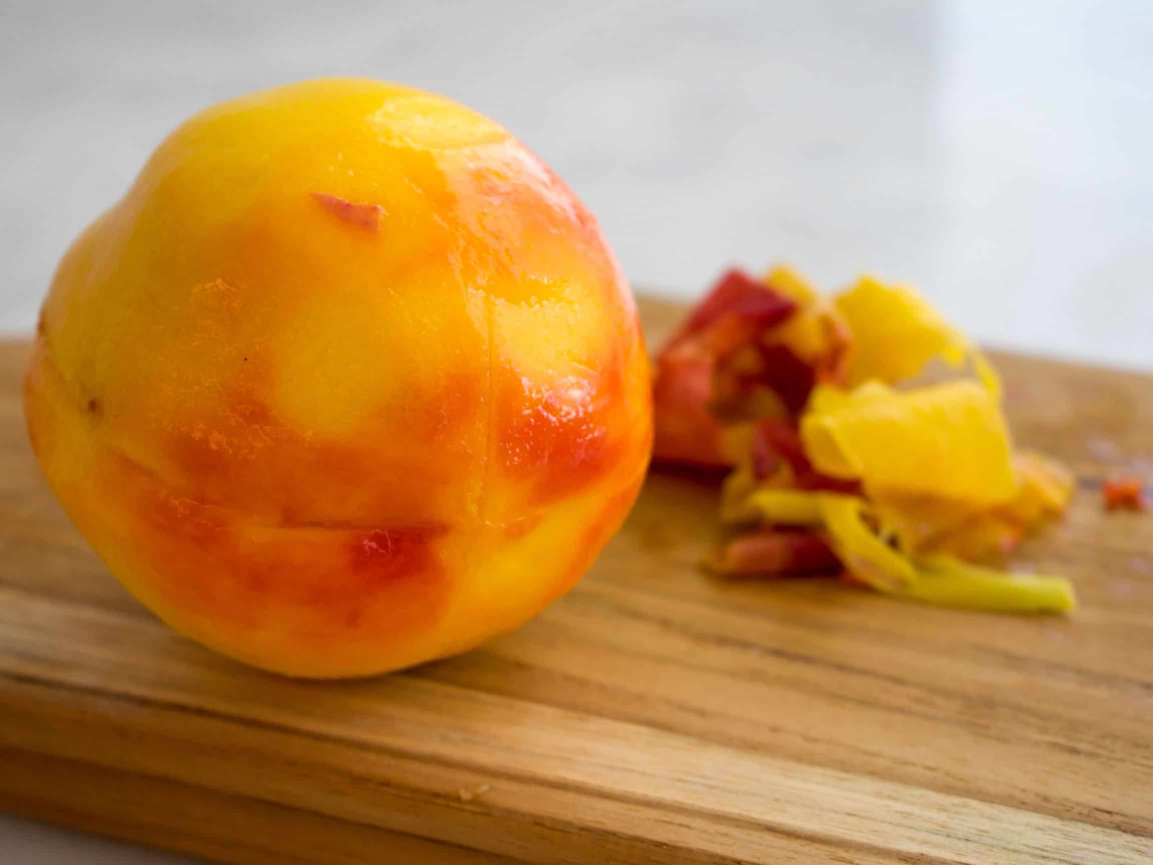 A blanched peach sitting on a wooden cutting board with the peels sitting beside.