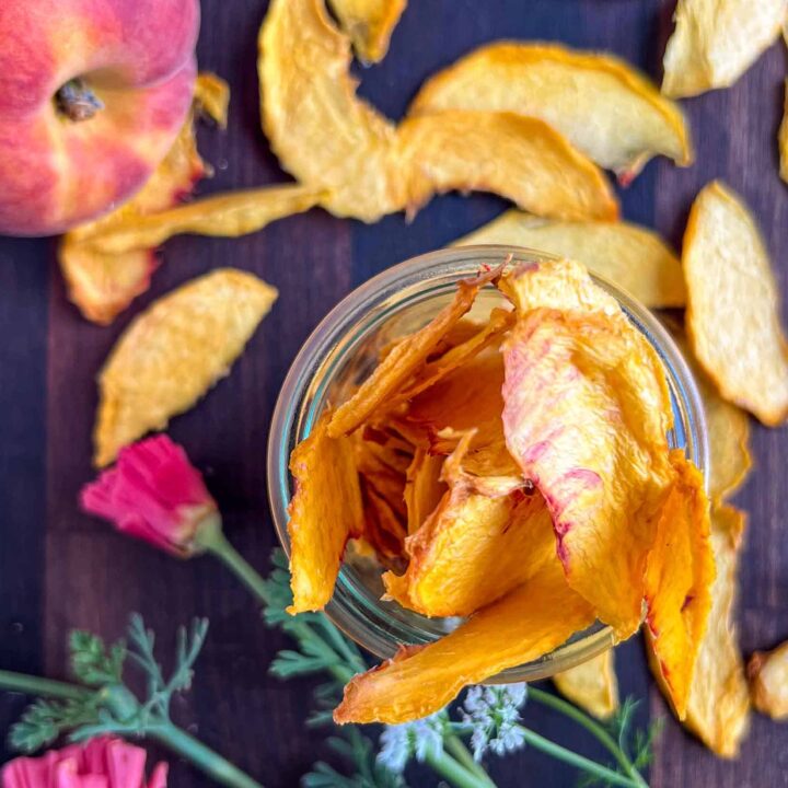 Dehydrated peaches piled high in a glass jar with dried peaches scattered around and a whole peach.