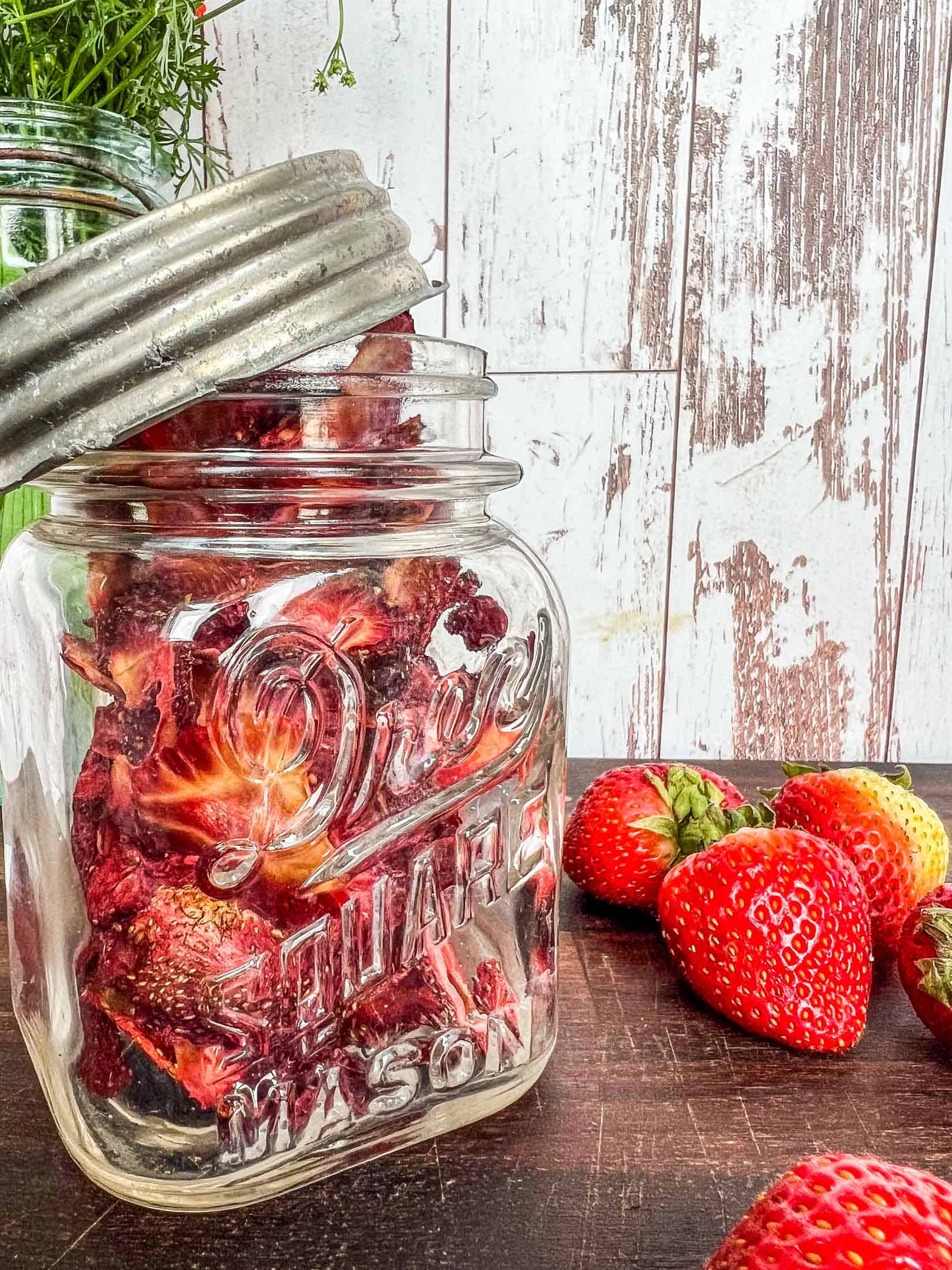 Dried strawberries in a decorative antique jar with a metal lid loosely hanging off of the rim.