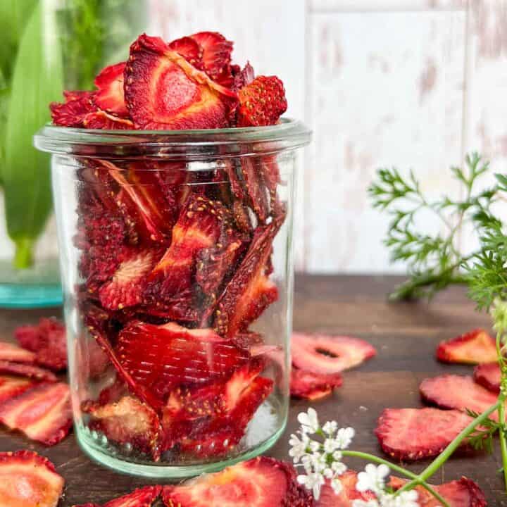 Dehydrated strawberries overflowing a small glass jar with dried slices around and some simple greenery beside.