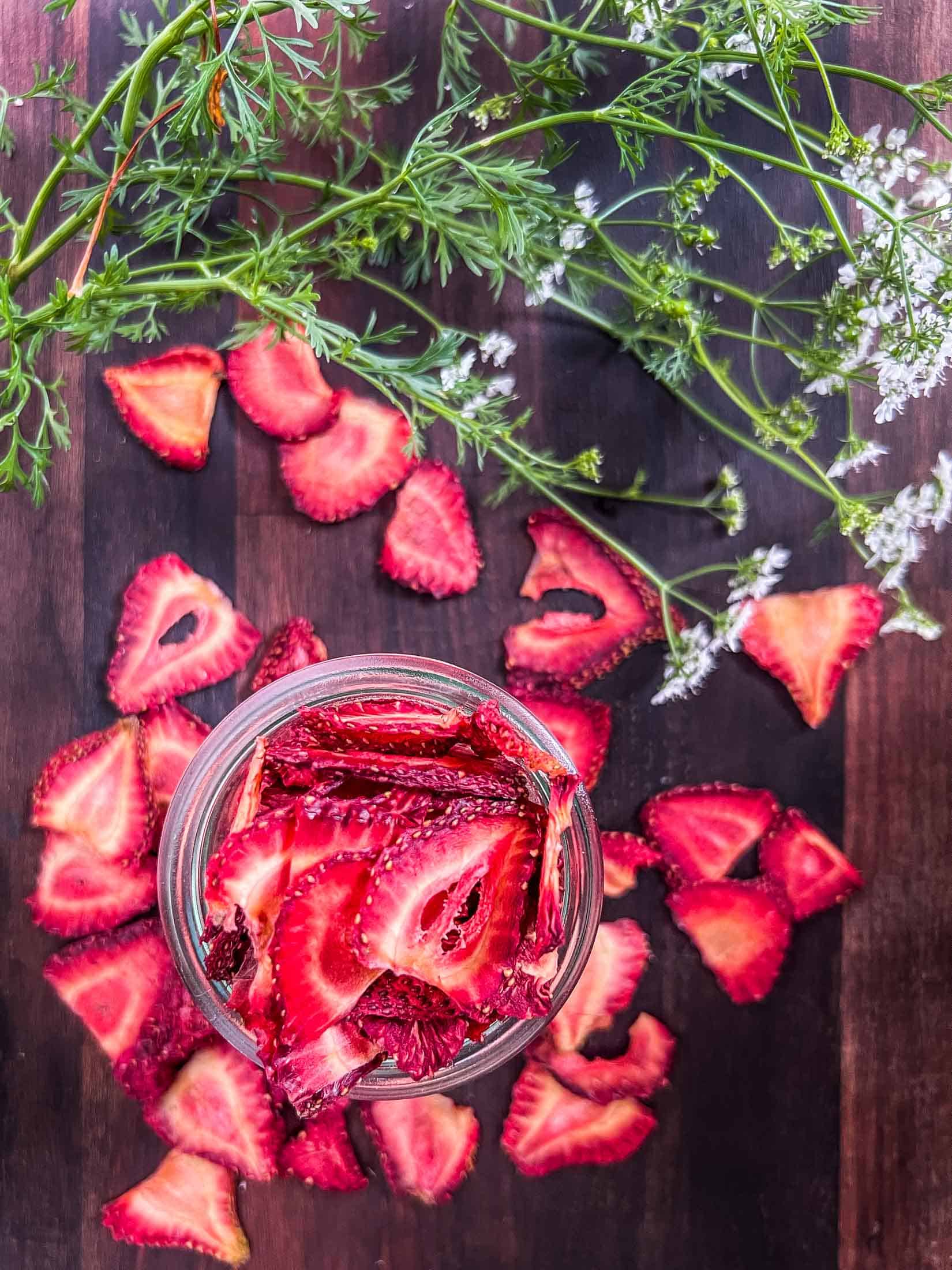 Dehydrated strawberries filling a glass jar with dried berries spilled out around and fresh greens beside.