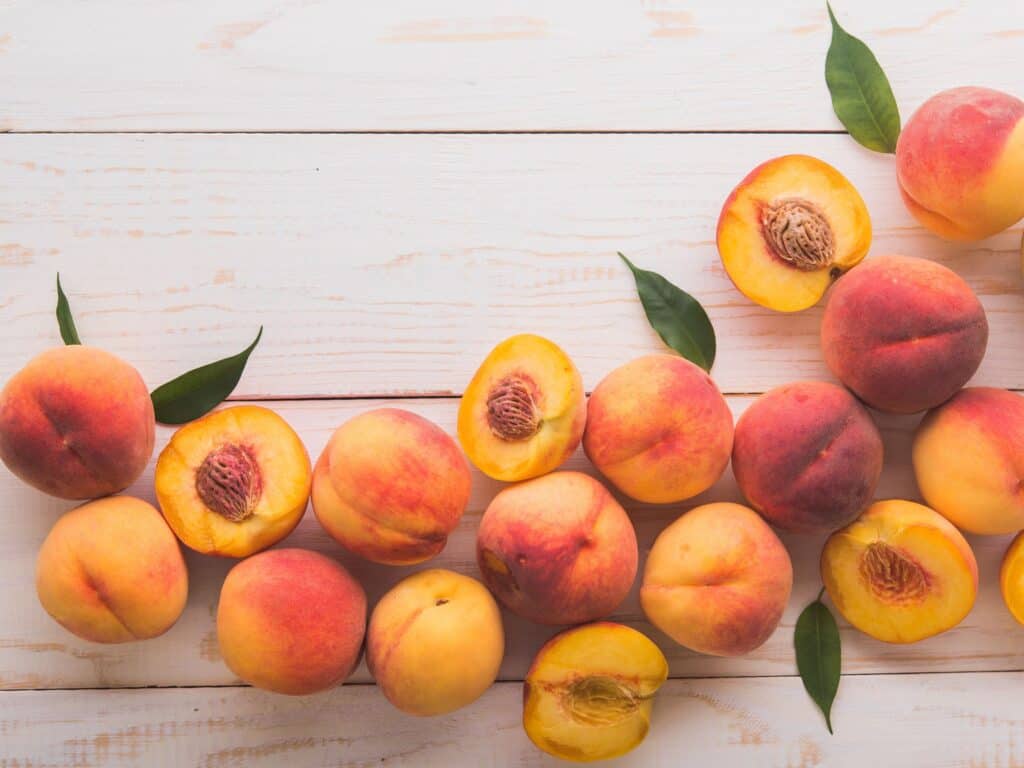 Whole and halved peaches on a wooden board with green peach leaves around.